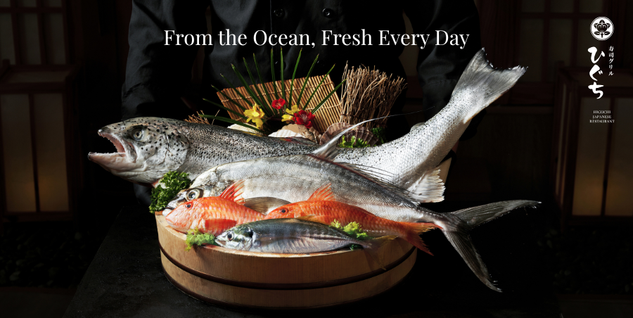 Fresh assorted whole fish on a wooden platter, garnished with greenery, set against a dark background for a seafood restaurant display.