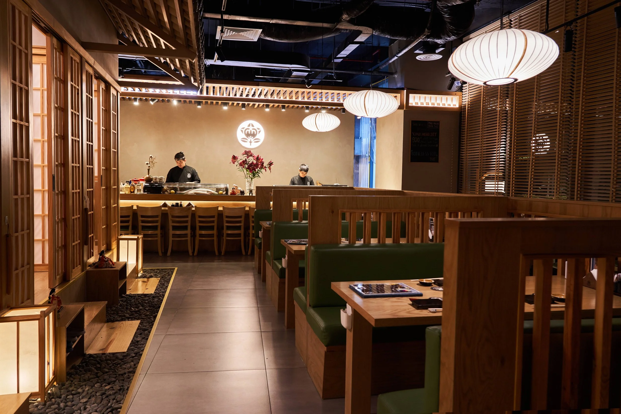 Interior of a modern Asian restaurant with wooden partitions, green cushioned seats, and hanging paper lanterns. Two chefs are working behind the counter, and there is a decorative flower arrangement on the counter. Warm lighting and traditional decor enhance the ambiance.
