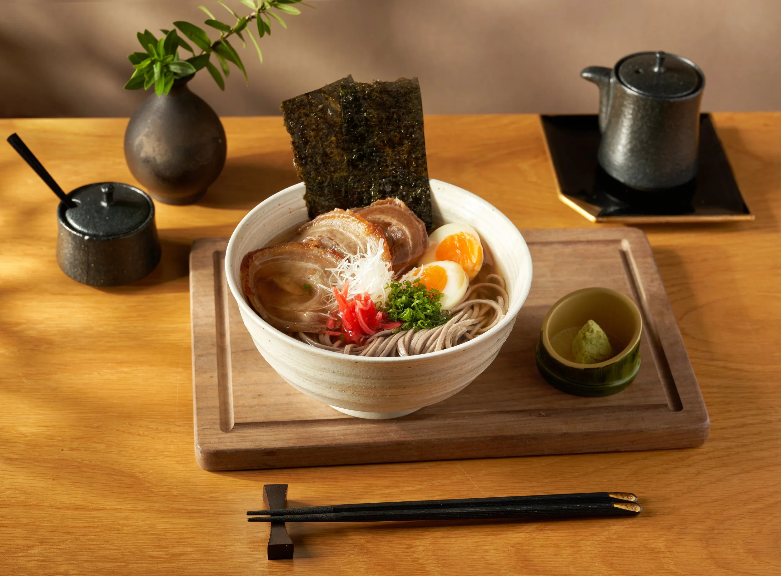 A bowl of ramen with sliced pork, half a boiled egg, shredded white radish, chopped green onions, and a sheet of nori on top. There are small side dishes with wasabi and other condiments, on a wooden tray with black chopsticks and two spice containers on a wooden table.