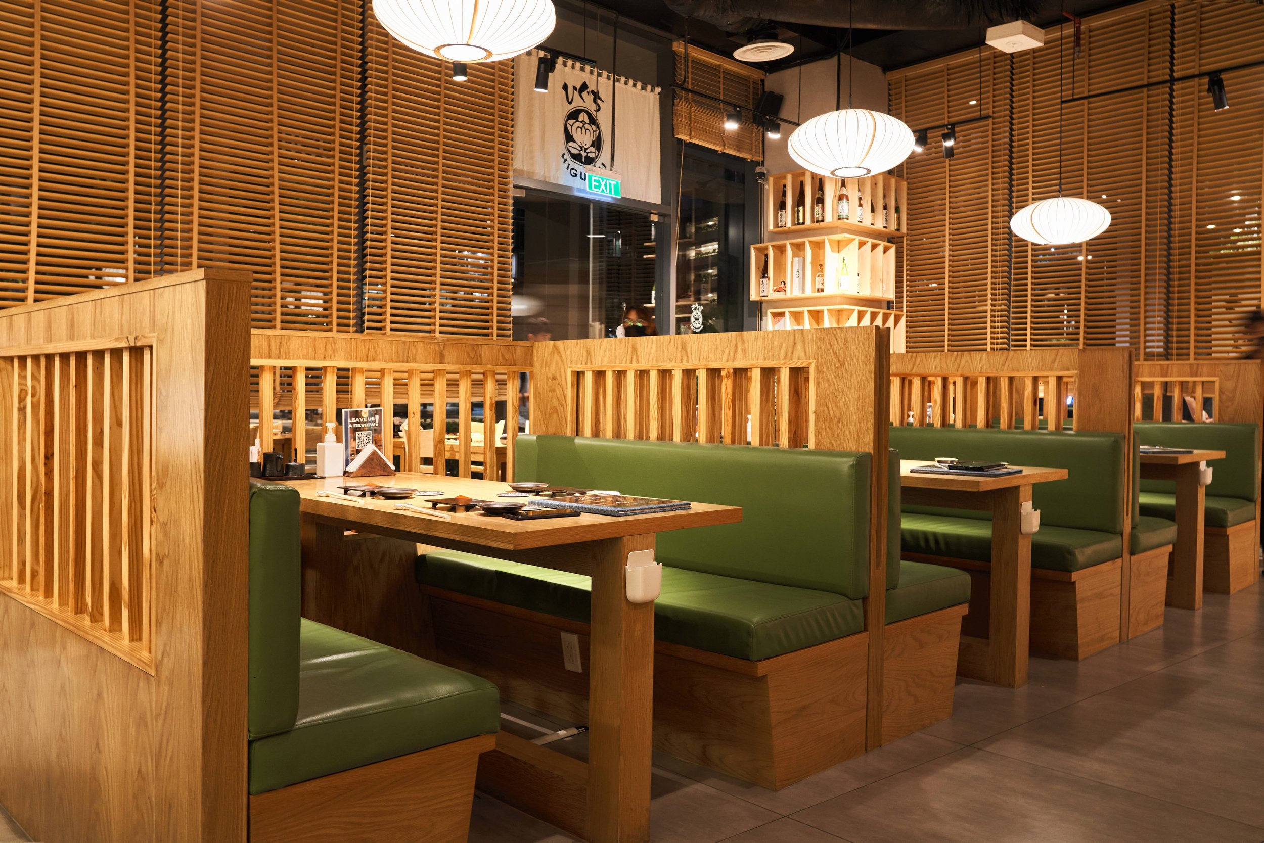 Interior of a Japanese restaurant with wooden partitions, green cushioned booths, hanging paper lanterns, and decorative wooden shelves.