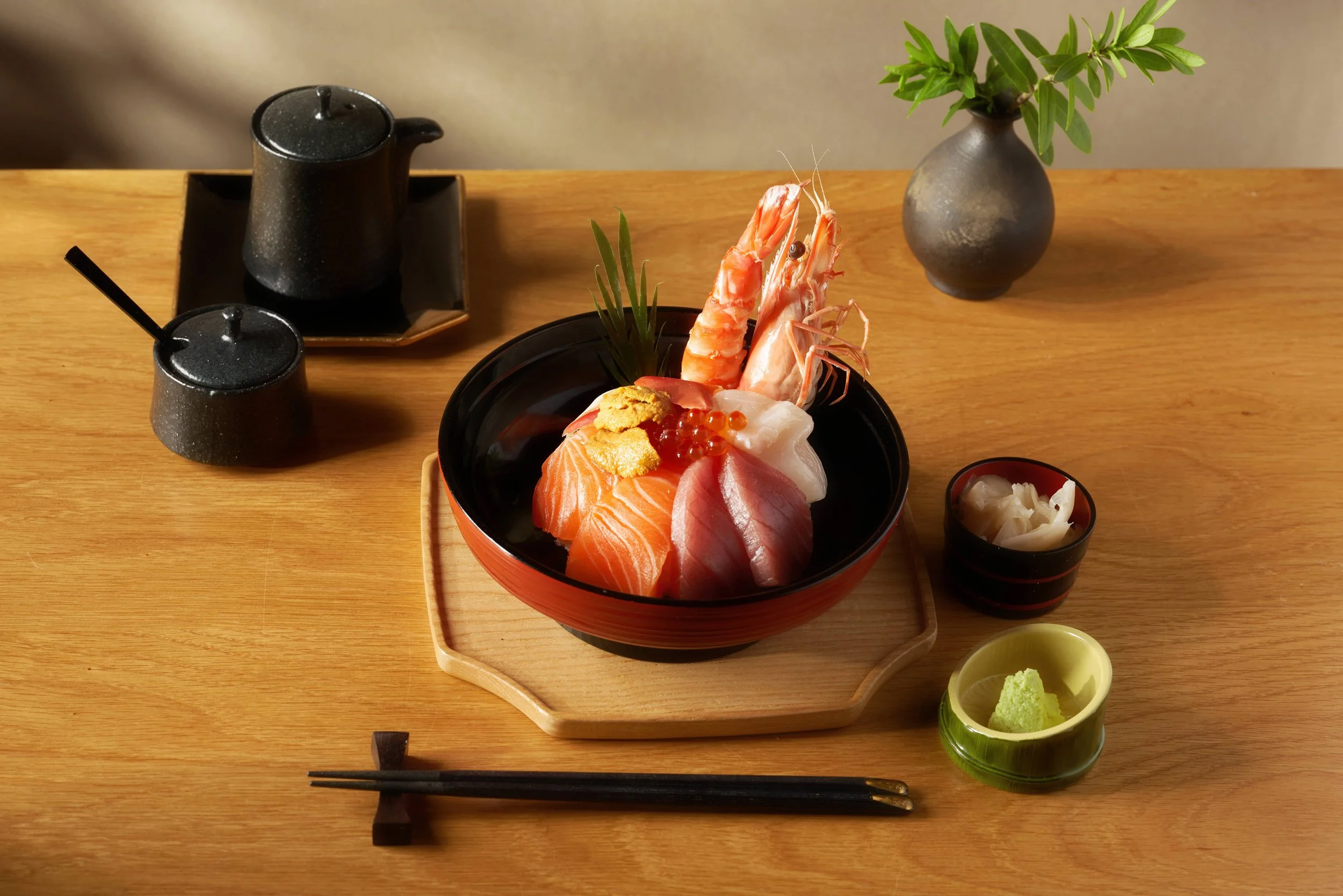 A bowl of assorted sashimi including salmon, tuna, shrimp, and eel, garnished with wasabi, served on a wooden tray with soy sauce, pickled ginger, and green leaves, set on a wooden table.