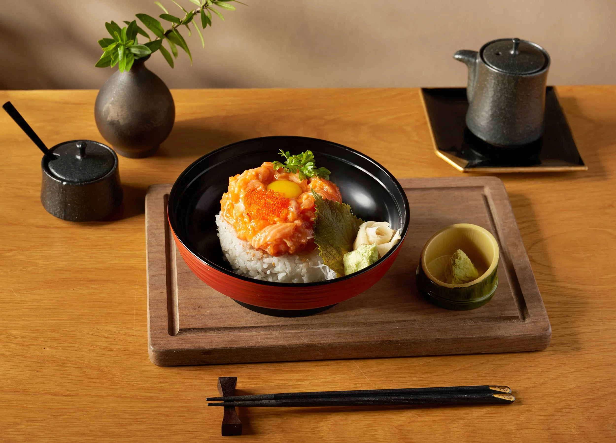 A Japanese meal featuring a bowl of rice topped with raw salmon, salmon roe, a raw egg yolk, and garnishes, served on a wooden tray with small dishes of wasabi, pickled ginger, and dipping sauce, alongside black chopsticks and a black cup, a gray teapot, and a small gray vase with green leaves.