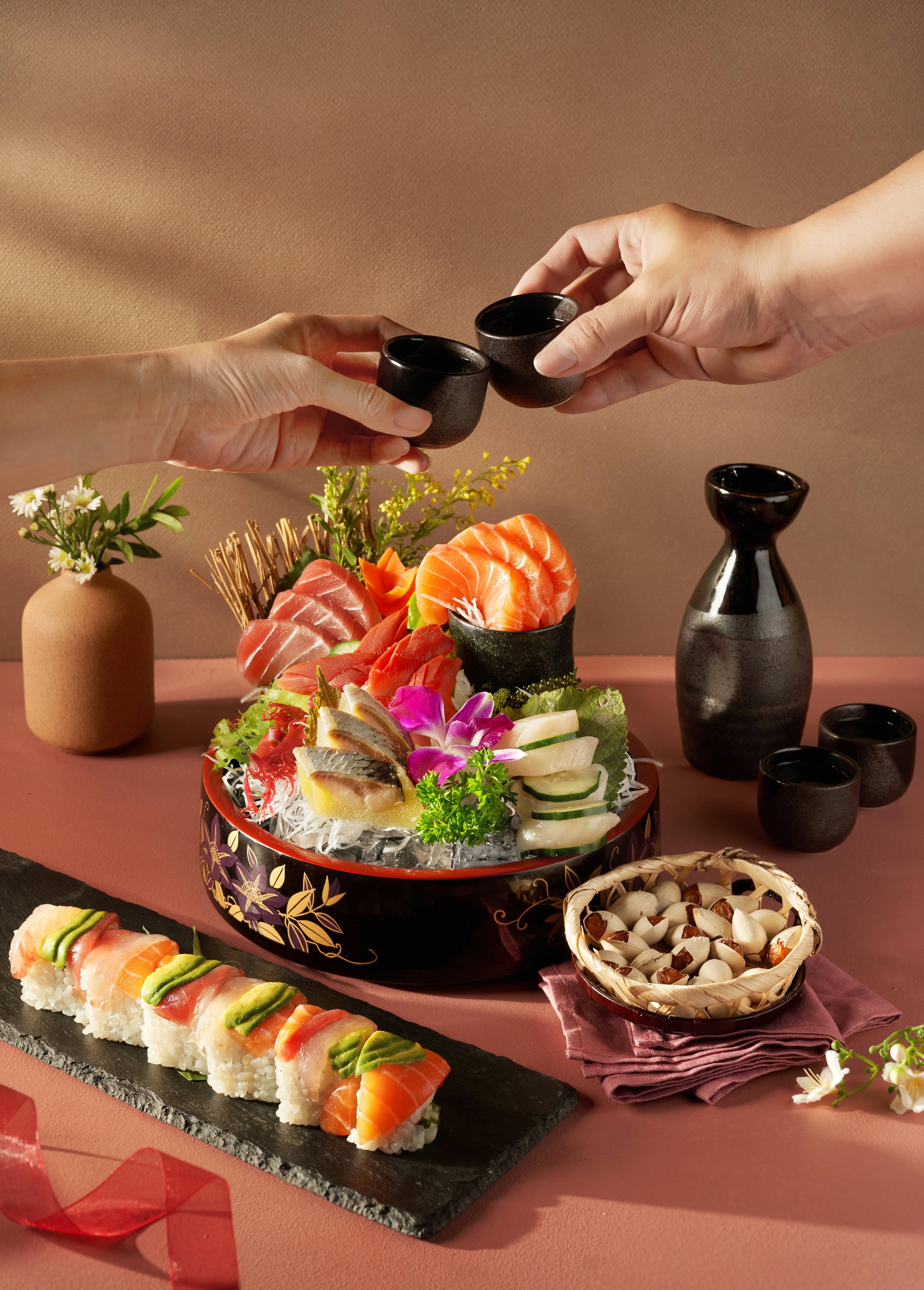Two hands exchanging small black ceramic cups. In the background, a platter of assorted sashimi and sushi on a black and gold tray, with various decorative elements, flowers, and a beige vase with flowers, all arranged on a pink surface.