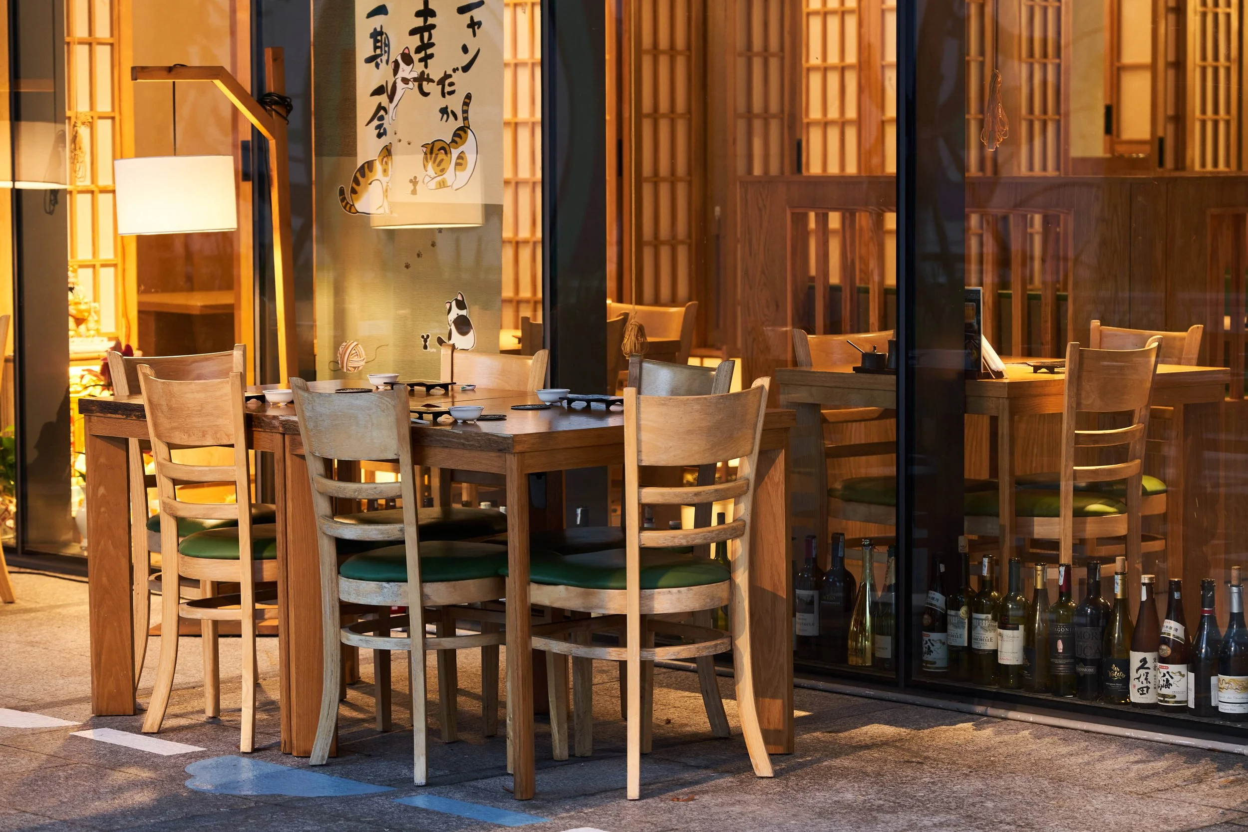 A wooden outdoor dining area with a table set with bowls and utensils, surrounded by wooden chairs, and a glass-front wine display with bottles inside, illuminated by warm lighting.