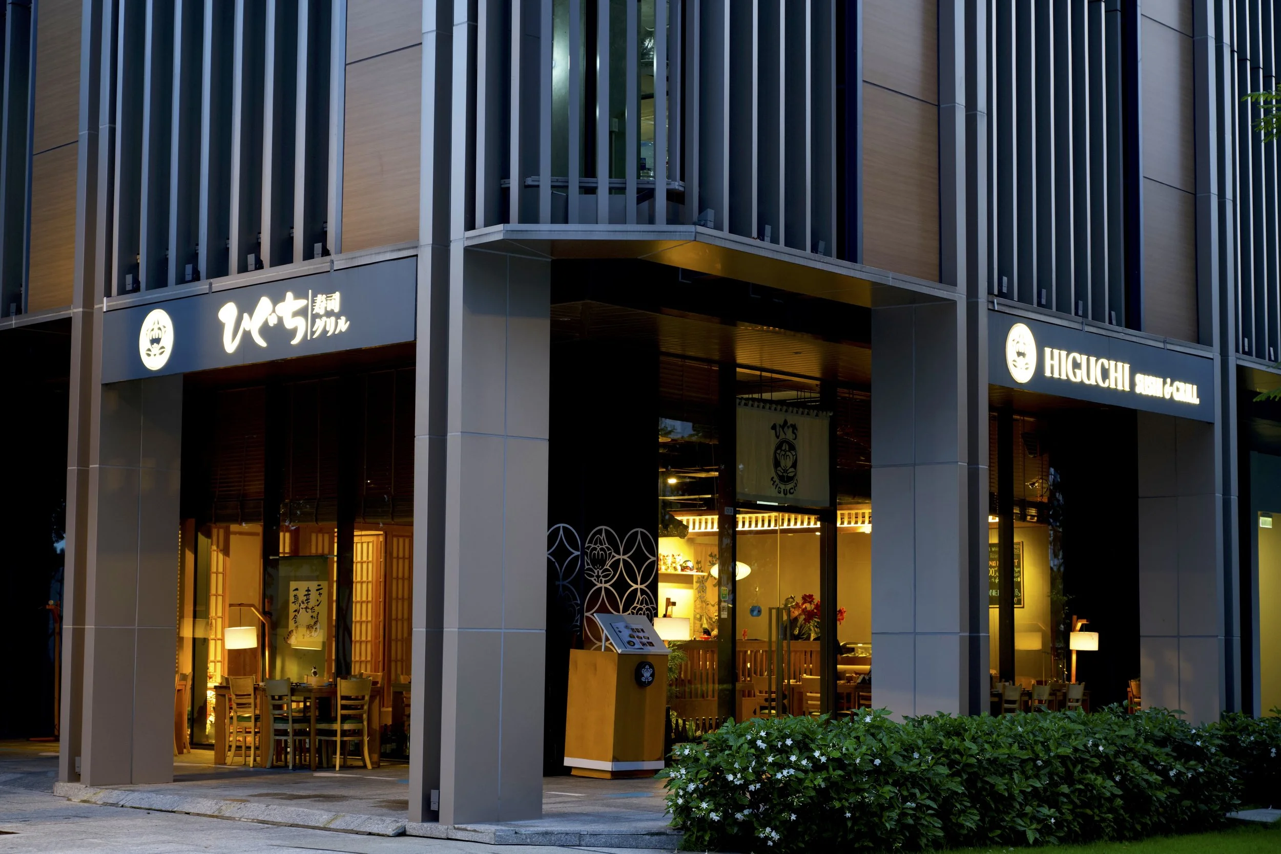 Exterior of a modern restaurant with illuminated sign in Japanese and English, large glass windows revealing interior dining area, and greenery outside.