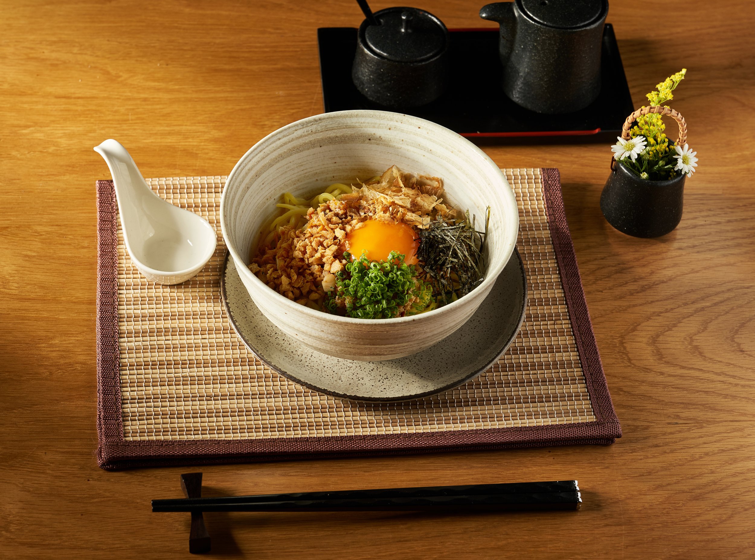 A bowl of Japanese udon noodles with a raw egg yolk, chopped green onions, shredded nori, and minced meat, placed on a red and beige woven placemat on a wooden table, accompanied by a small white spoon and a black tea set with a teapot and cups in the background, and a small black vase with yellow and white flowers.
