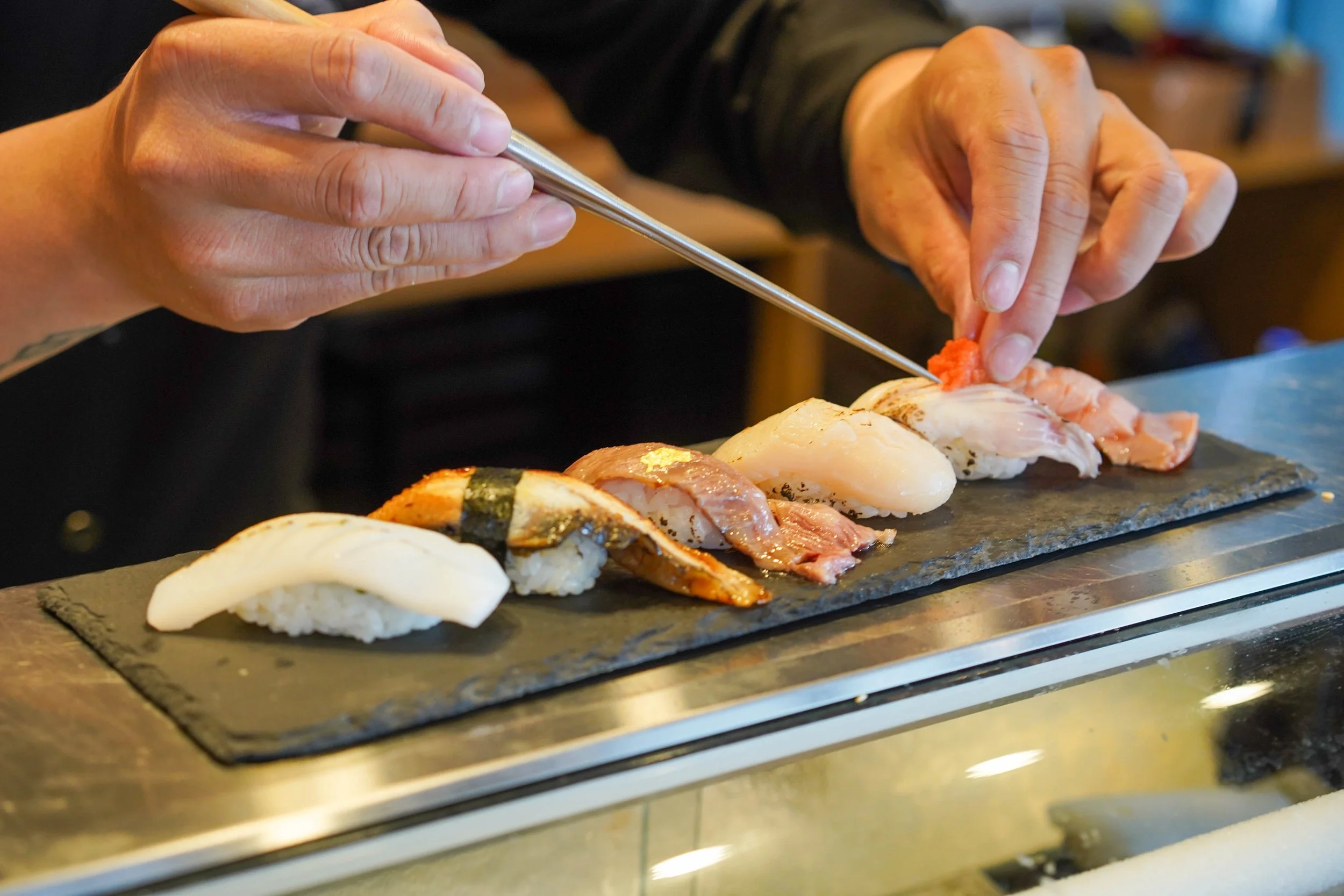 Close-up of a person using chopsticks to pick up pieces of sushi on a black slate platter, with assorted sushi pieces including fish, shrimp, and eel on a table.