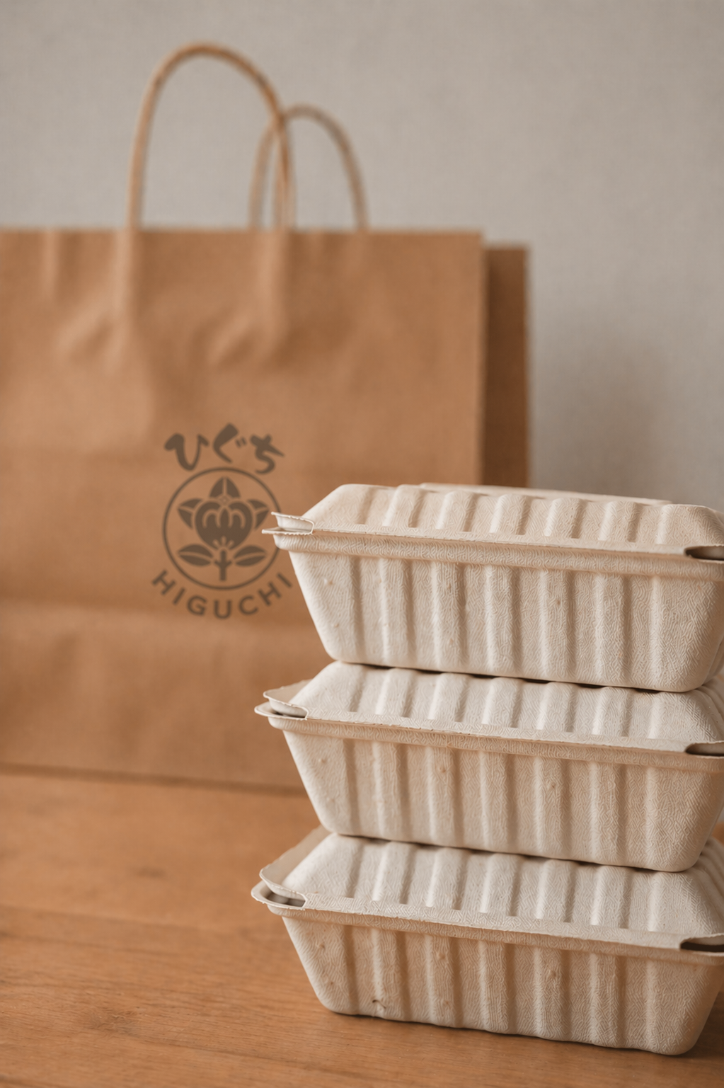 Three stacked beige takeout containers in front of a brown paper bag with Japanese characters and a floral logo.