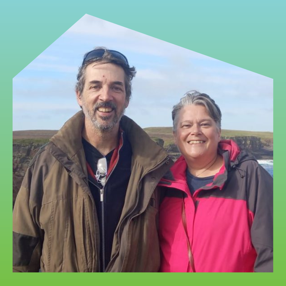 A smiling man and woman standing outdoors on a cloudy day with a grassy cliff in the background.