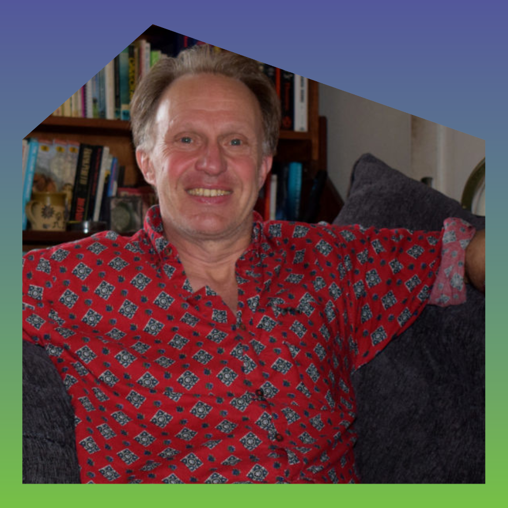 A middle-aged man with light skin, light brown hair, and a wide smile, wearing a red patterned shirt, sitting on a couch in a room with bookshelves in the background.