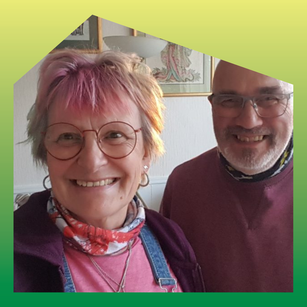 A smiling woman with pink hair, wearing glasses, earrings, a pink shirt, a patterned scarf, and a purple jacket, standing next to a smiling man with glasses, a beard, wearing a maroon shirt and a neck gaiter, in a room with framed artwork on the wall.