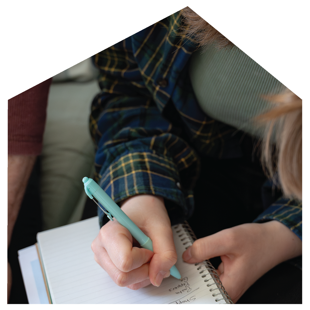 A person wearing a plaid shirt sitting on a couch and writing in a notebook with a pastel green pen. Their head is slightly bent down, partially visible with light-colored hair. Part of another person's arm and a sleeve are also visible in the background.