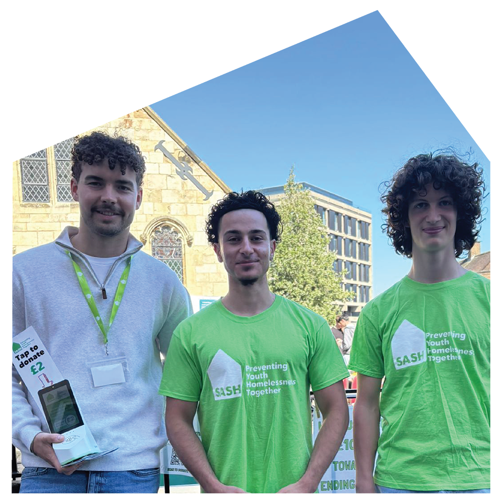 Three young men standing outdoors during daytime, wearing green T-shirts with the SASH logo and the slogan 'Preventing Youth Homelessness Together.' The man on the left is holding a donation box and has a lanyard around his neck. The background features a historic stone building with windows and a modern office building, with trees and a clear blue sky.