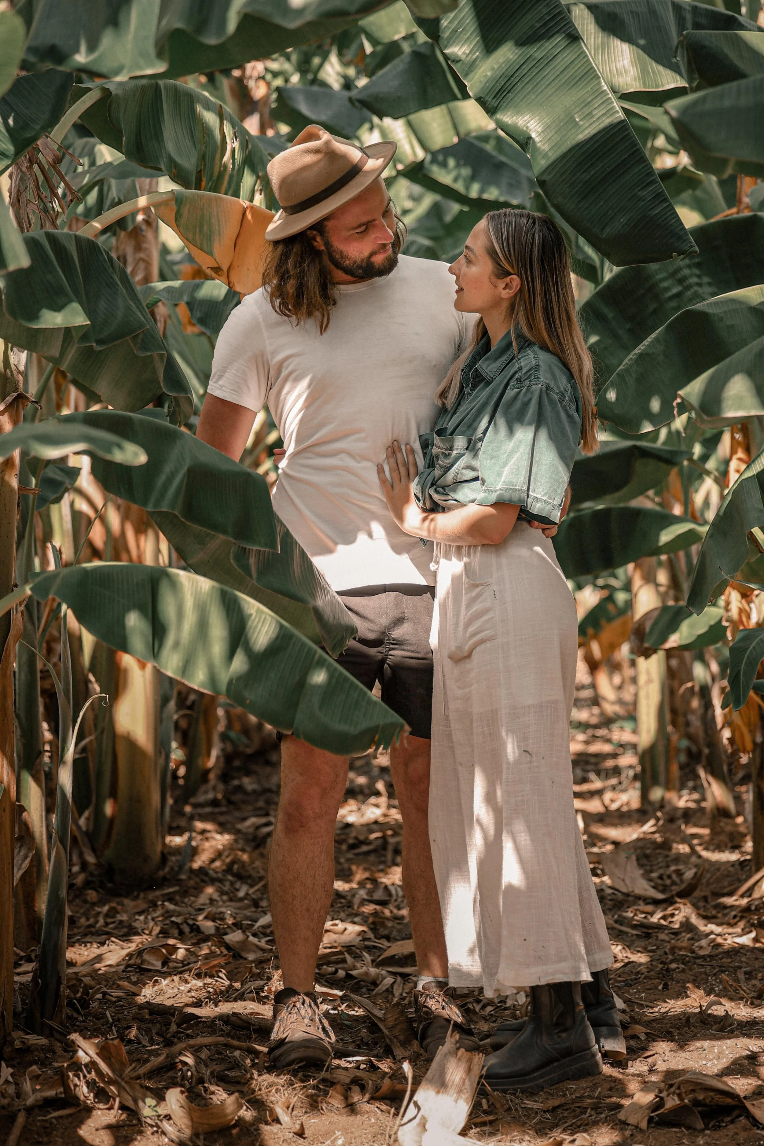 A couple standing close together among tall green banana plants, looking into each other's eyes in a forest setting.