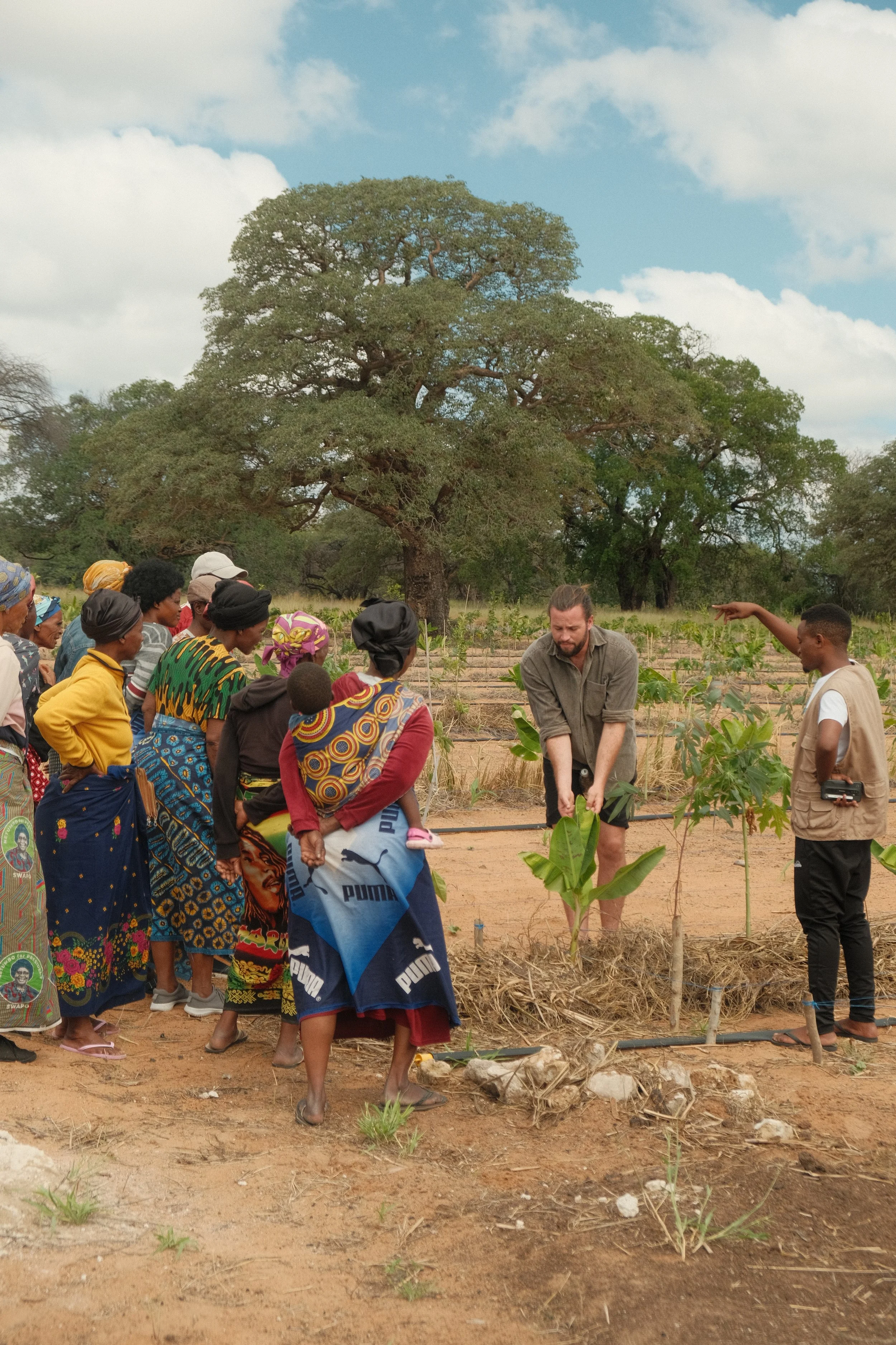 A group of people, including women and children, gathered outdoors on a farm or rural area, listening to a man with a beard and gray shirt. The background features a large tree, green plants, and a cloudy sky.