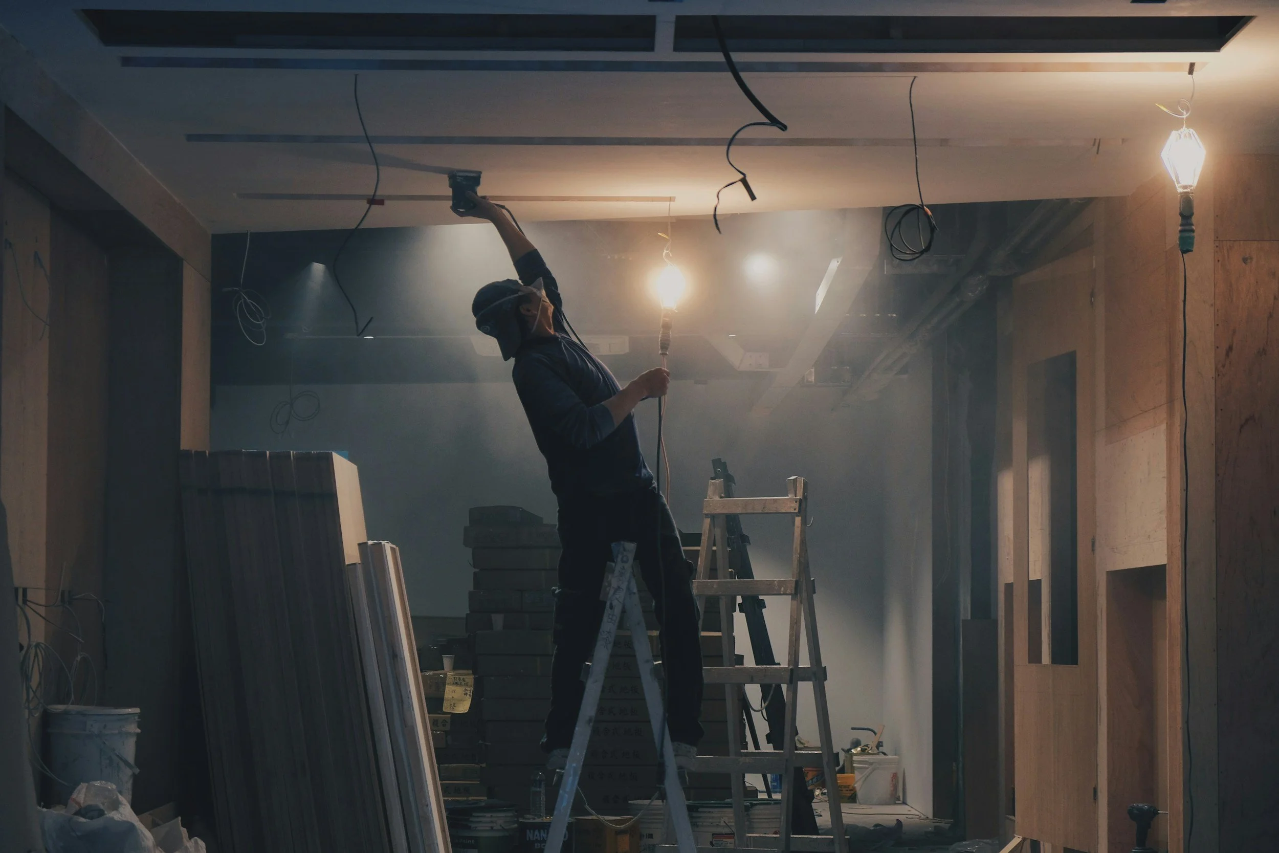 A construction worker standing on a ladder, installing or wiring electrical fixtures on a ceiling in a building under construction with construction materials and tools around.