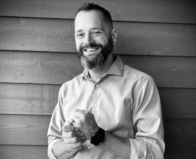 A smiling man with a beard, wearing a button-up shirt and a watch, standing against a wooden wall.