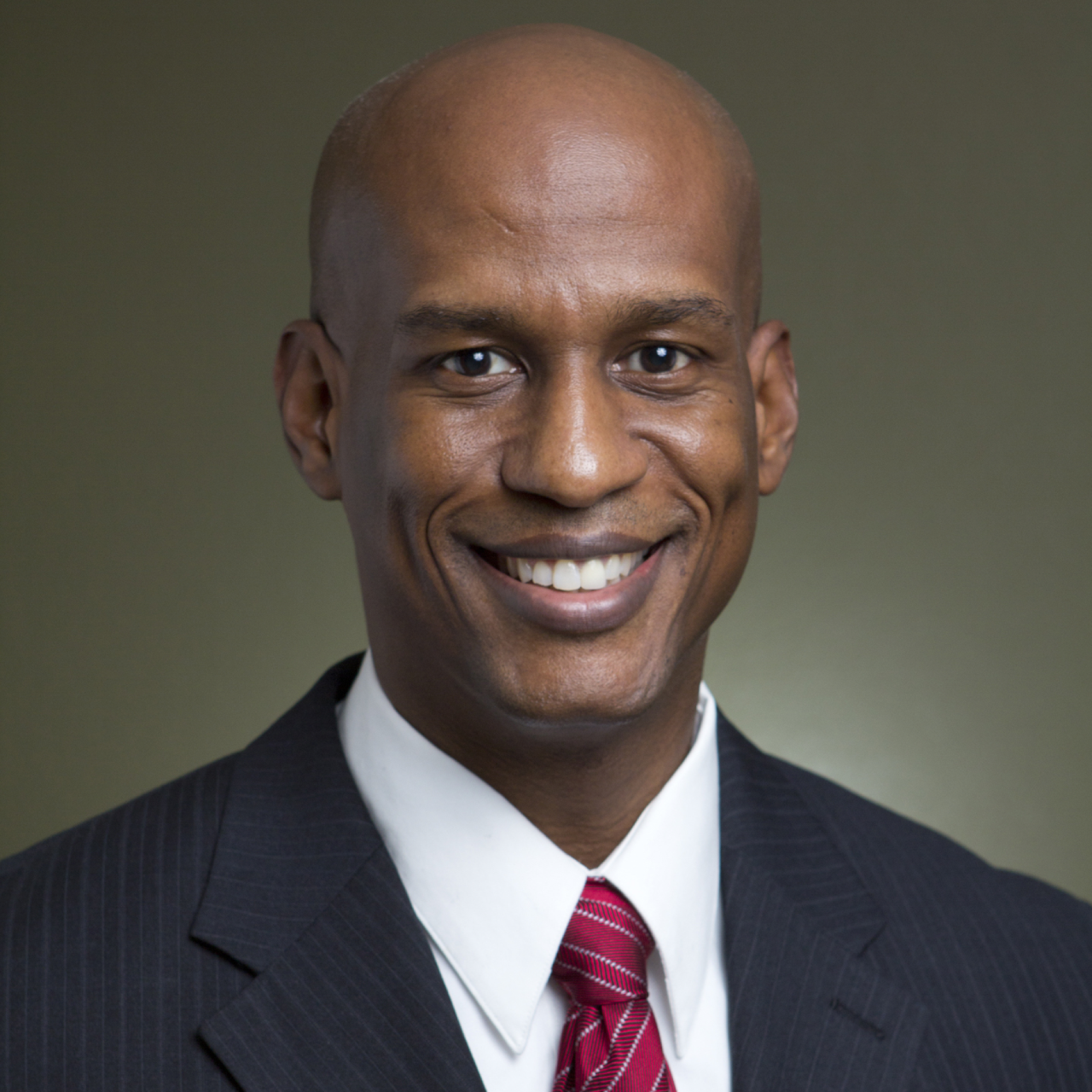 Professional portrait of a smiling Black man in a business suit, white shirt, and red tie against a plain background.