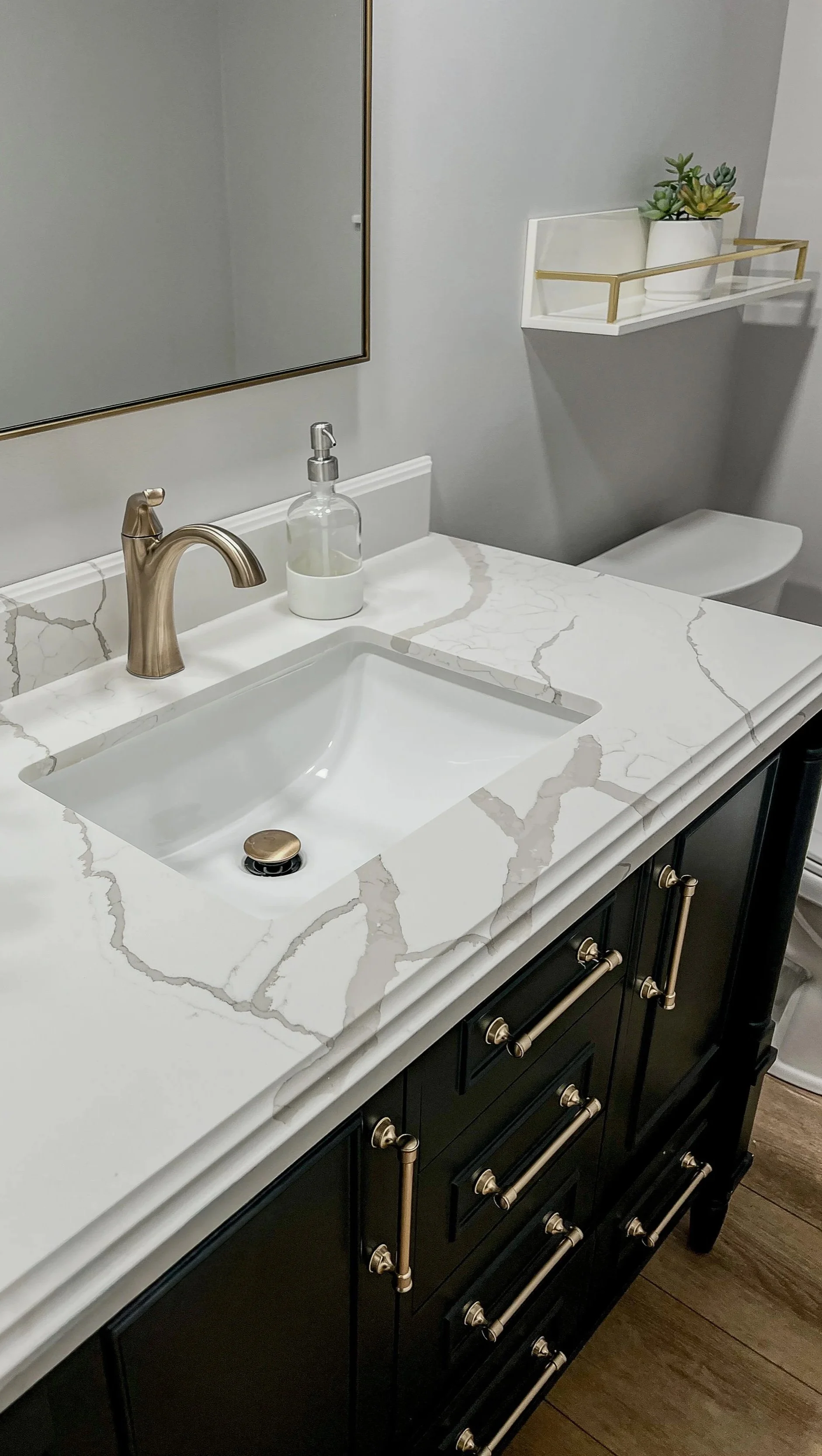 Dark bathroom vanity with a quartz countertop, brass faucet, and brass wall shelf in a Centerville remodel.