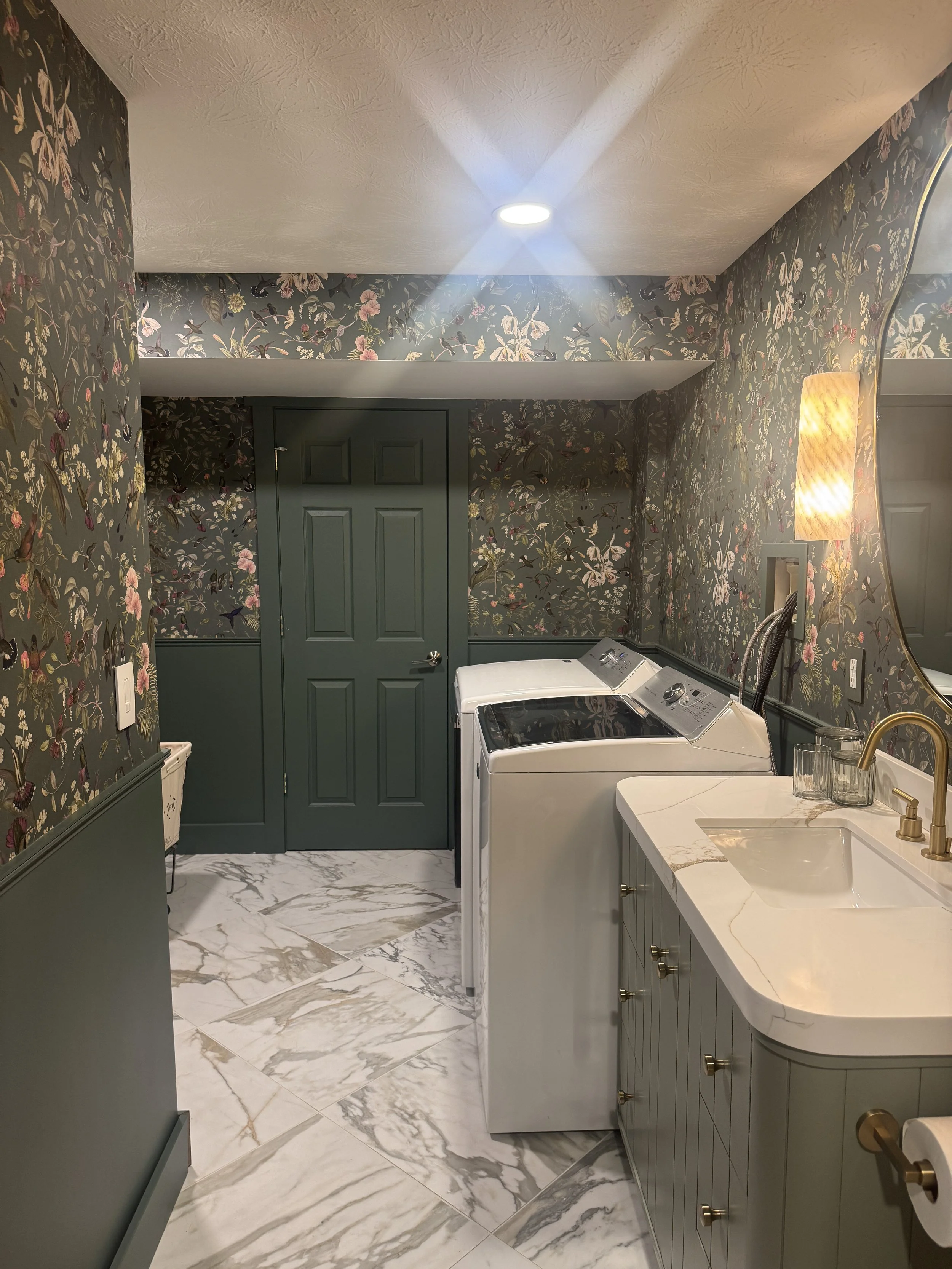 Full view of a Beavercreek, Ohio basement bathroom and laundry room renovation with side-by-side washer and dryer, marble floors, sage vanity, and botanical wallpaper by White Oak Interiors