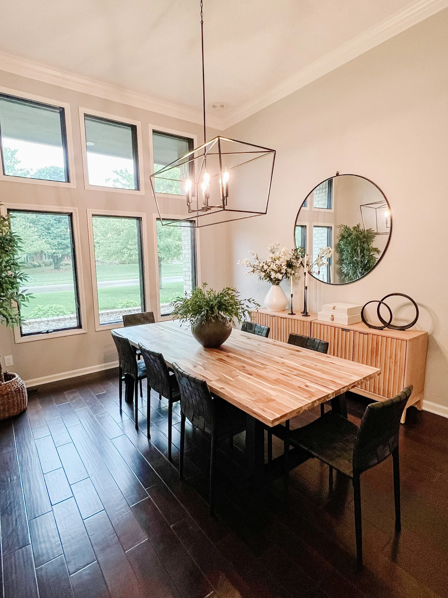 Dining room with a wall of windows, a wood table with black chairs, a linear chandelier, and a wood sideboard with a round mirror in a Centerville, Ohio home