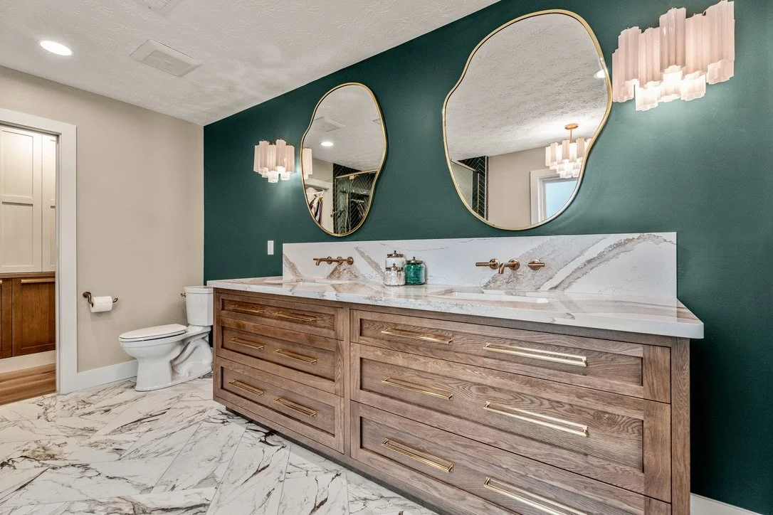 Wood double vanity with veined quartz countertop, wall-mounted gold faucets, two organic-shaped mirrors, and a green accent wall in a Xenia, Ohio master bathroom