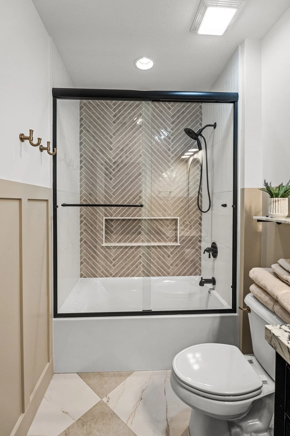 Glass-enclosed tub and shower combination with herringbone tile, matte black fixtures, and board-and-batten wainscoting in a Centerville, Ohio basement bathroom.