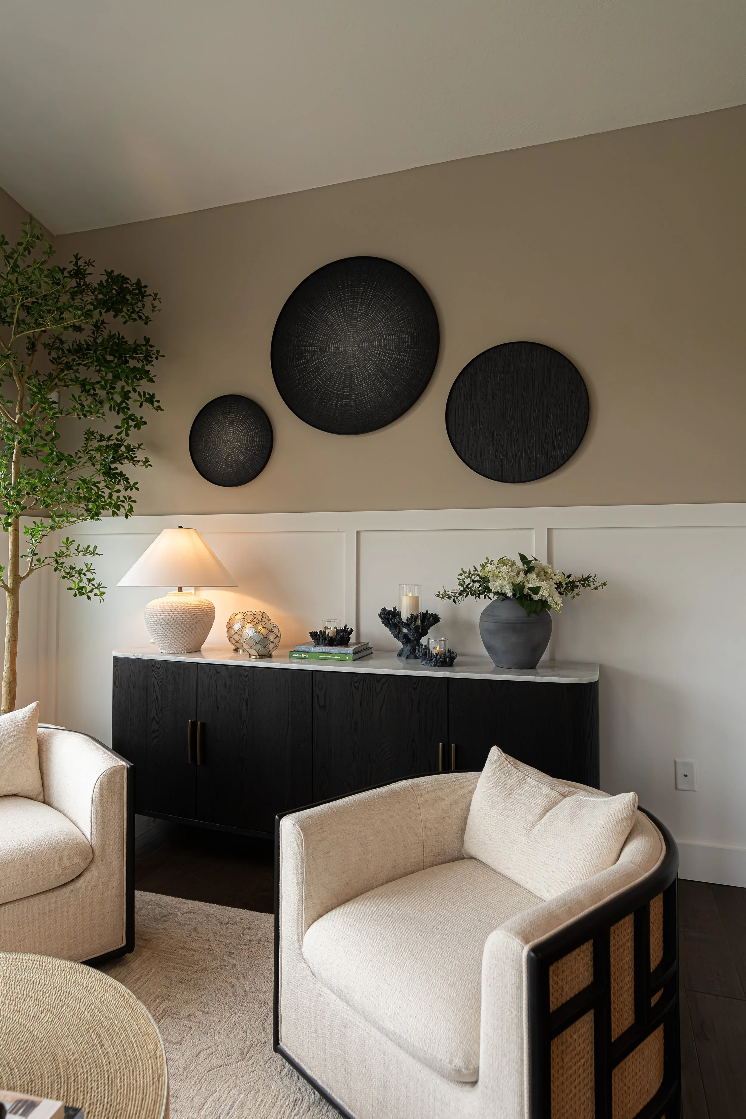 Sitting room with a black console, ceramic lamp, and grouping of woven wall discs above cream and cane chairs in a Centerville home renovation.