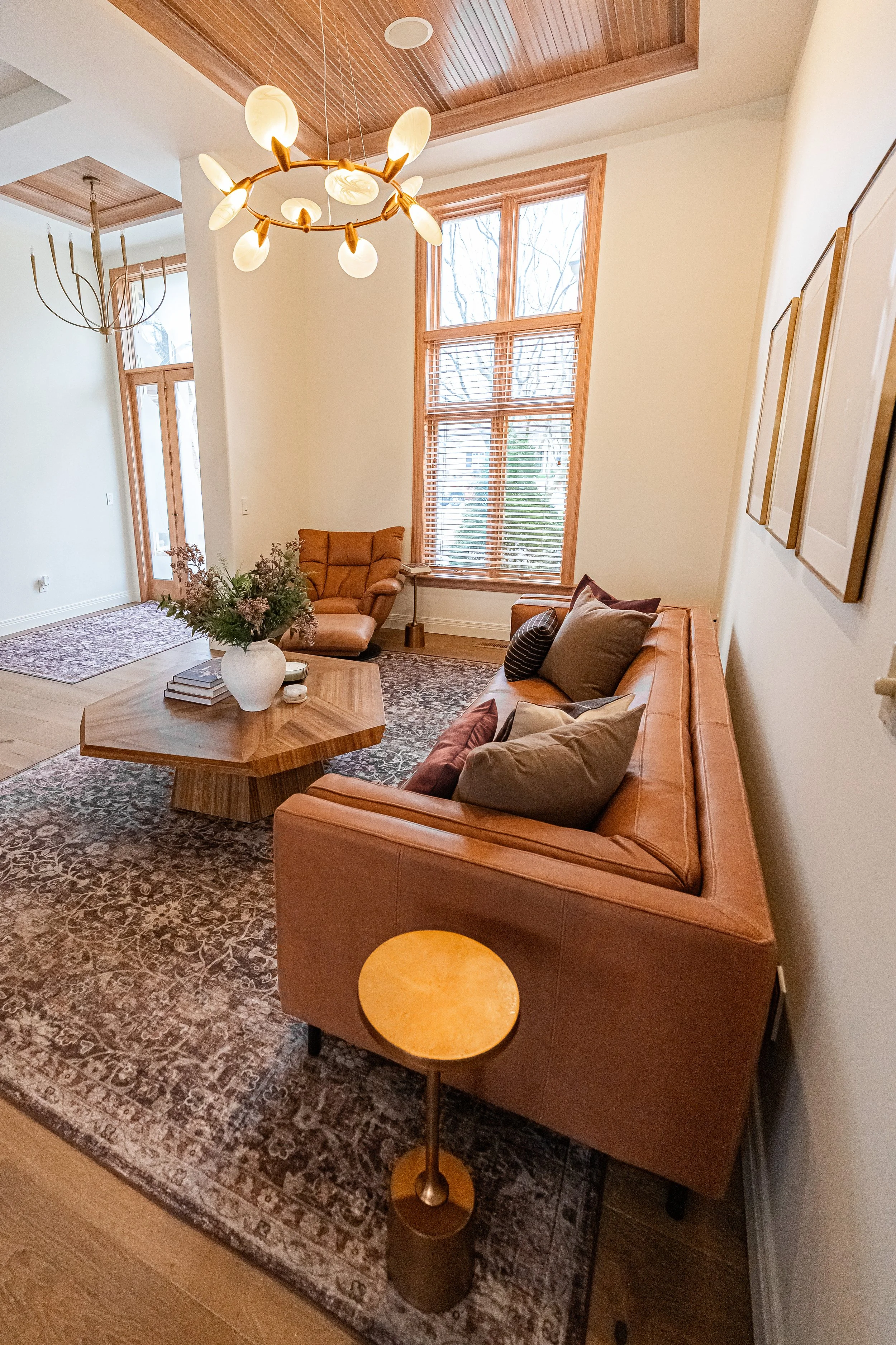 Living room with large wooden framed windows, tan leather sofa with throw pillows, patterned area rug, wooden coffee table with flower arrangement, side table with books, decorative table lamp, and pendant light fixtures hanging from wood-paneled cei