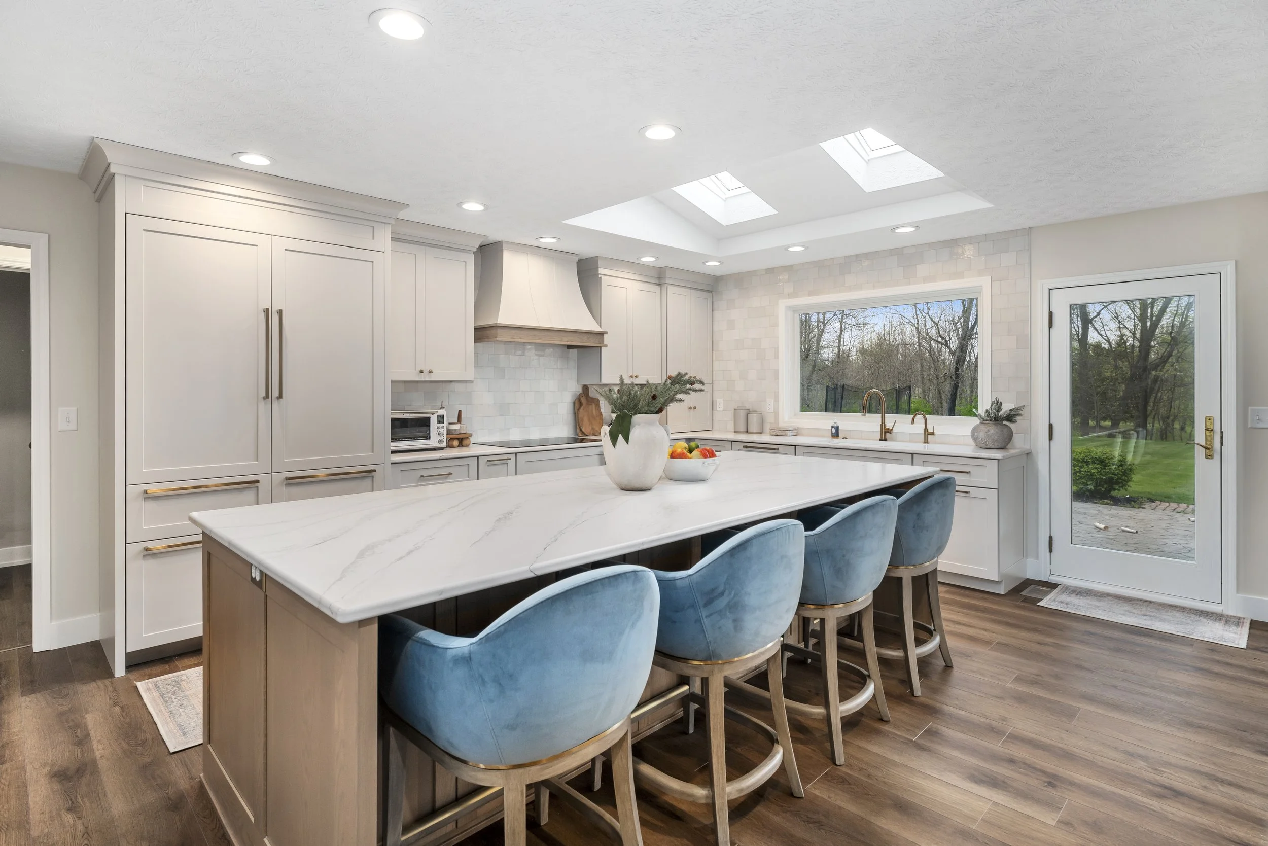 Renovated kitchen with light wood island, white quartz waterfall countertop, blue velvet barstools, custom hood, and a large window over the sink