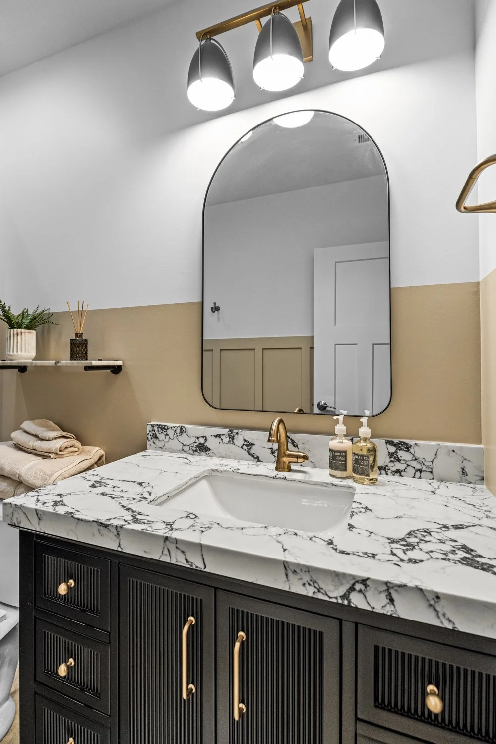Arched frameless mirror above a dark reeded vanity with a white-and-black marble countertop and brass faucet in a Centerville, Ohio basement bathroom.
