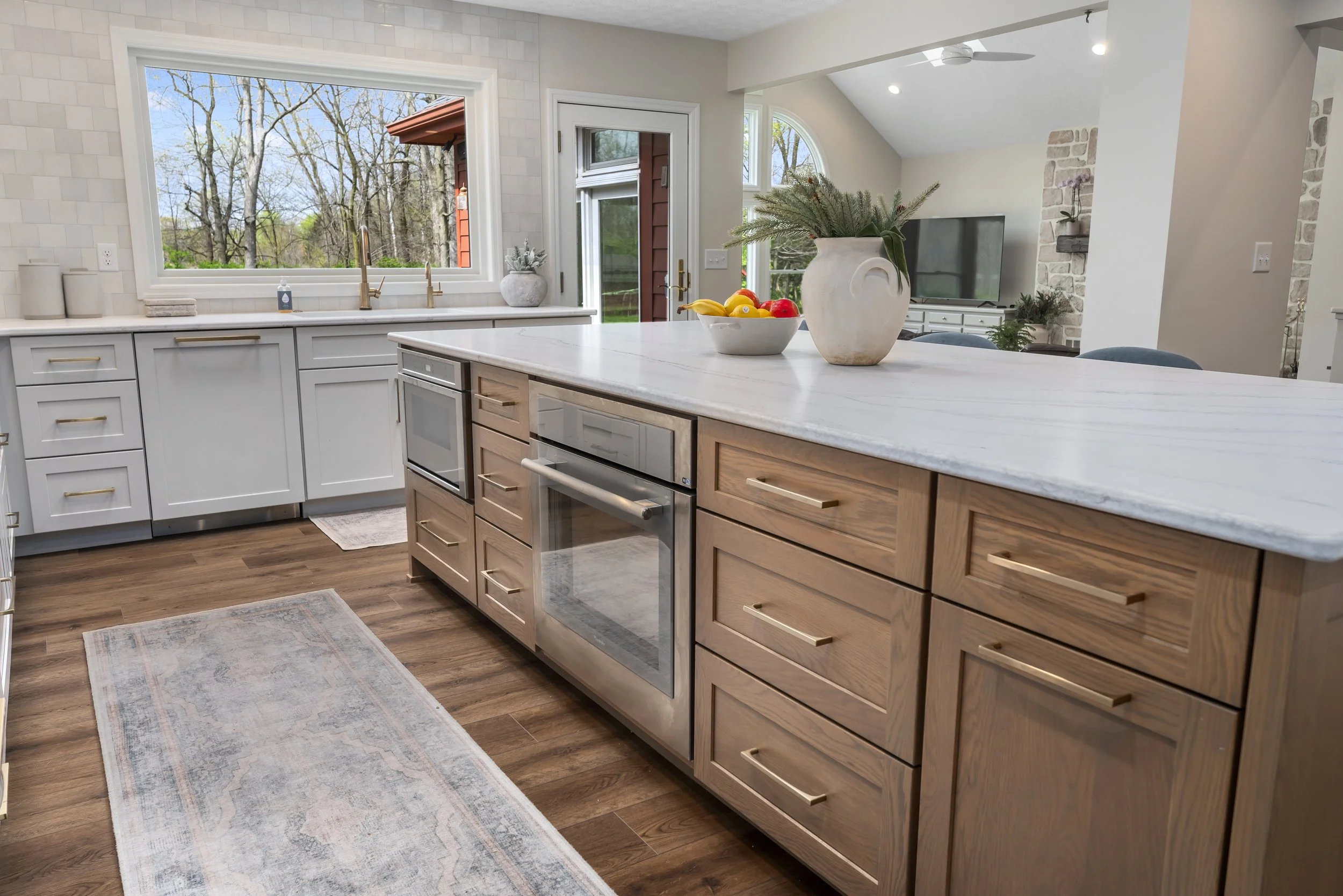 Light wood kitchen island with built-in oven and white quartz countertop, opening to the living room with natural light from multiple windows