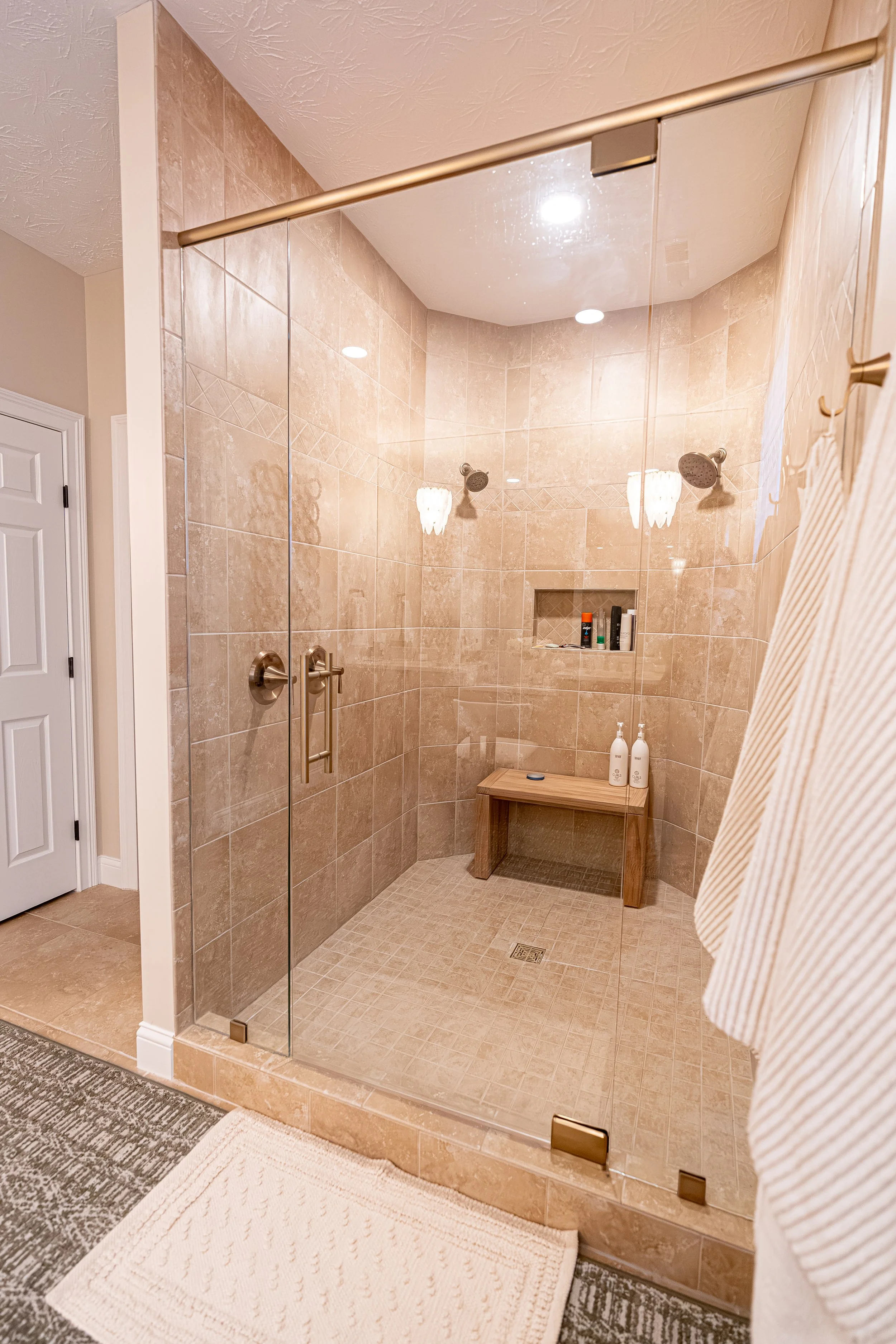 Enlarged glass-enclosed master shower with stone-look tile, built-in niche, teak bench, and brushed gold fixtures in an Oakwood, Ohio home
