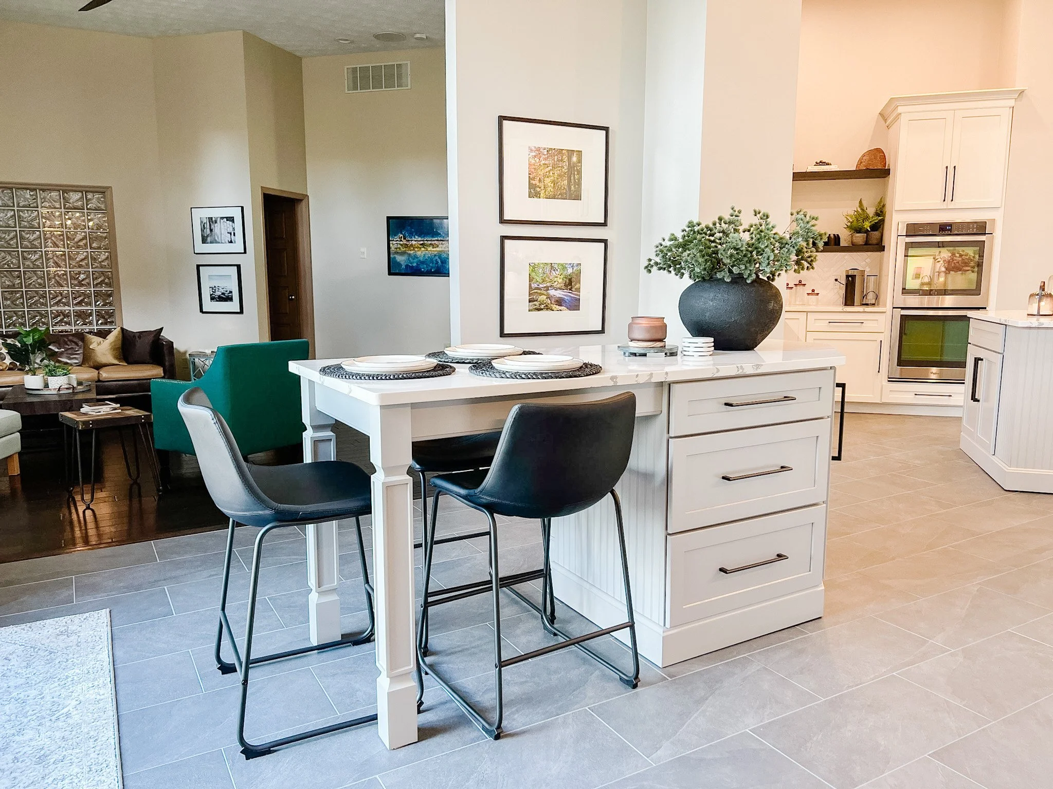 Open eat-in area with an island table and stools, a framed gallery wall, green accent seating, and the kitchen beyond in a Centerville, Ohio home
