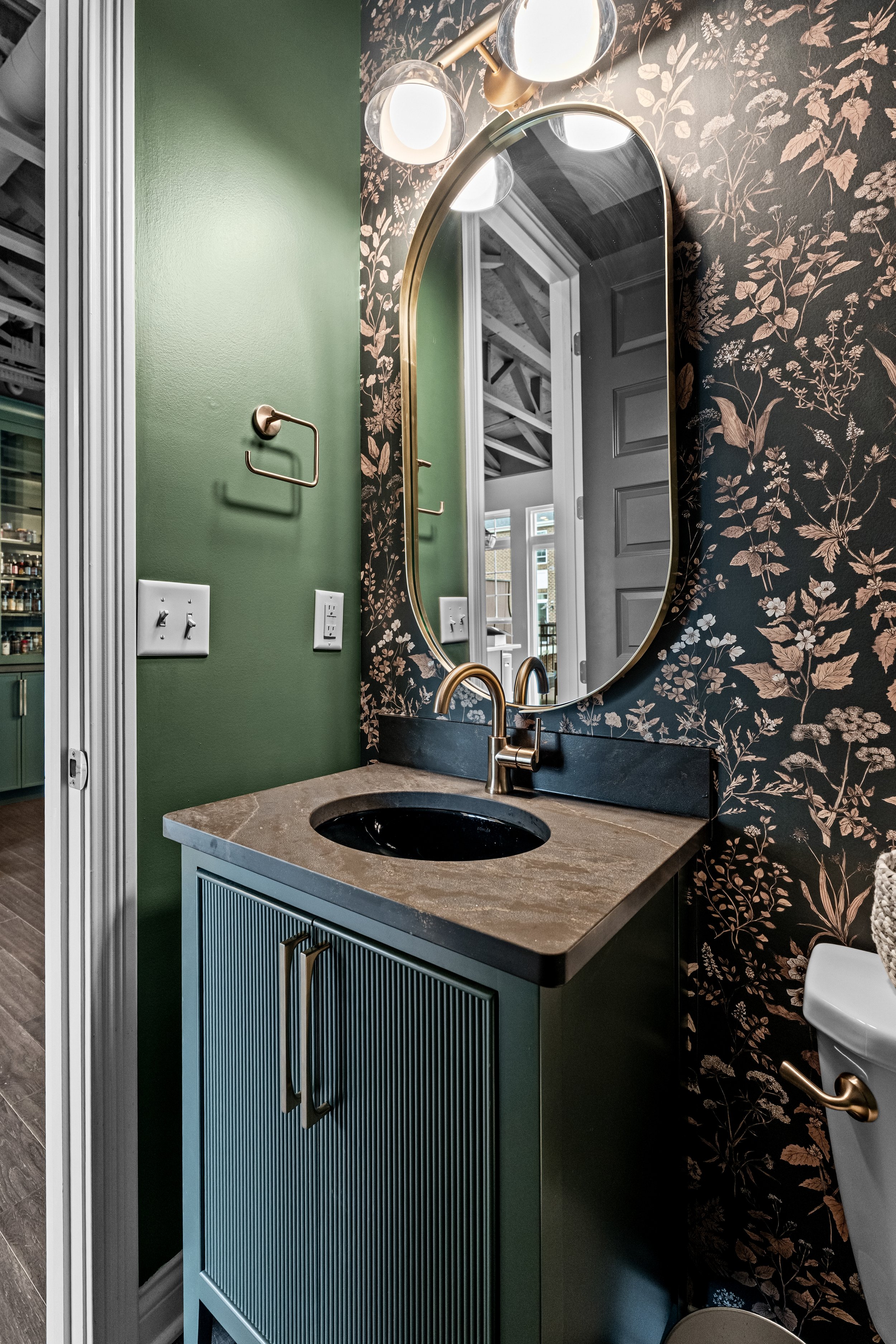 Remodeled half bath in Springboro, Ohio with a gold mirror, floral wallpaper, green reeded vanity, and smoky stone countertop by White Oak Interiors