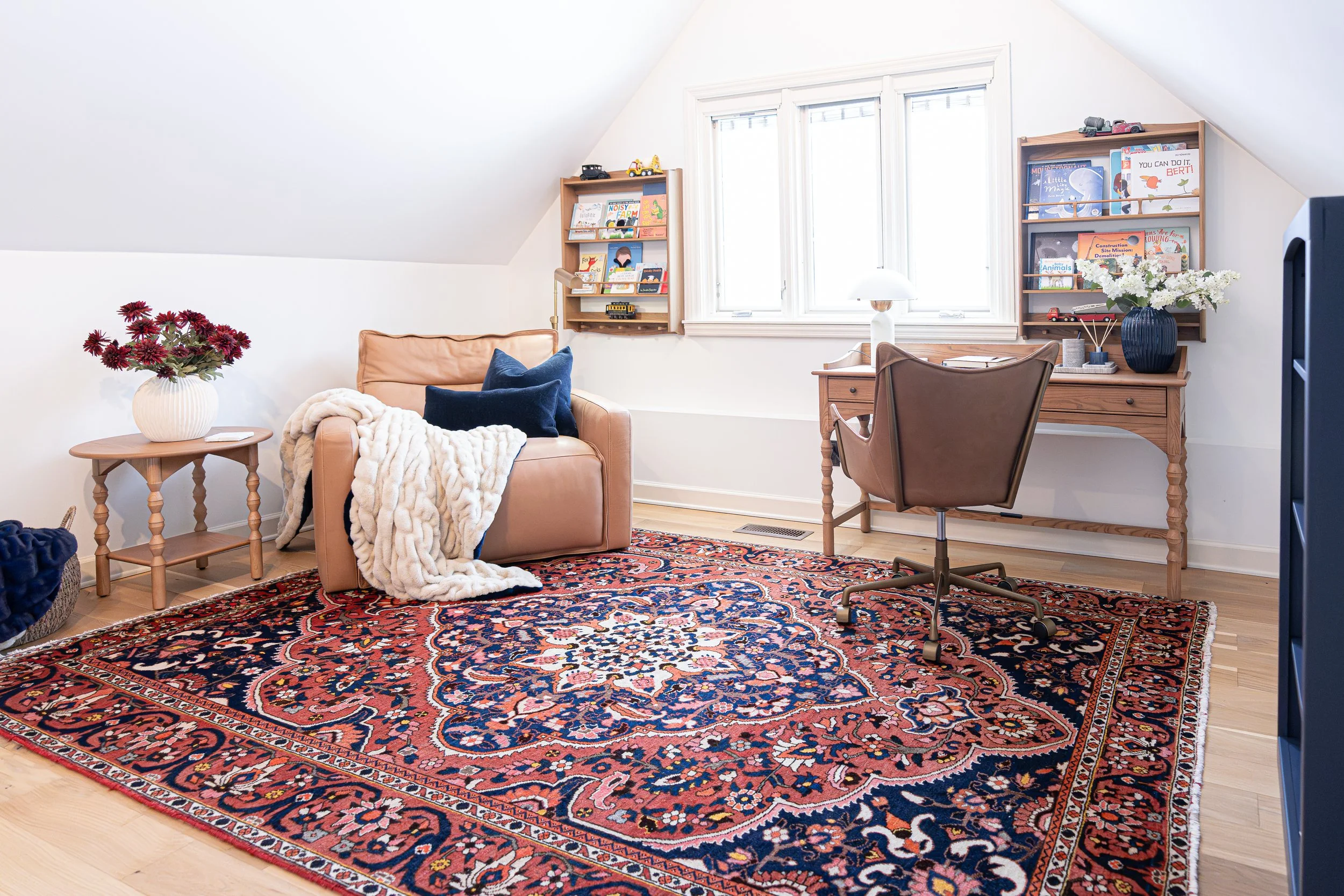 A cozy living room with a large, ornate red and navy oriental rug, a tan leather armchair with navy pillows and a white blanket, a small wooden side table with a white vase of red flowers, a wooden desk with a brown leather chair, paintings and books on wall shelves, a window with white frames, and a white table lamp.