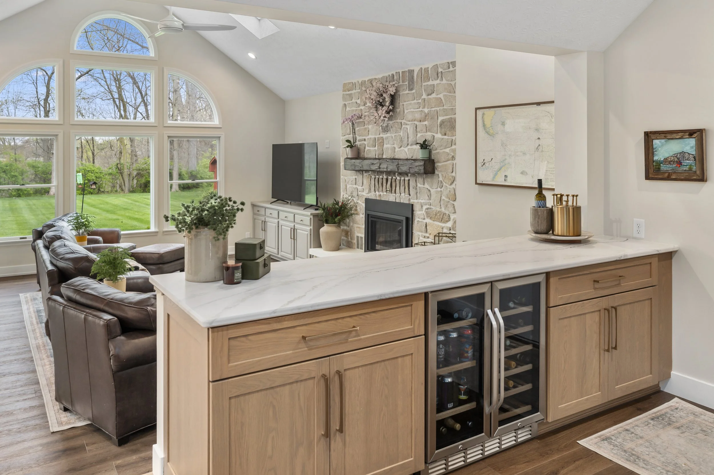 Open living room with a custom wood bar, quartz countertop, built-in beverage fridges, leather seating, and a stone fireplace beneath vaulted windows