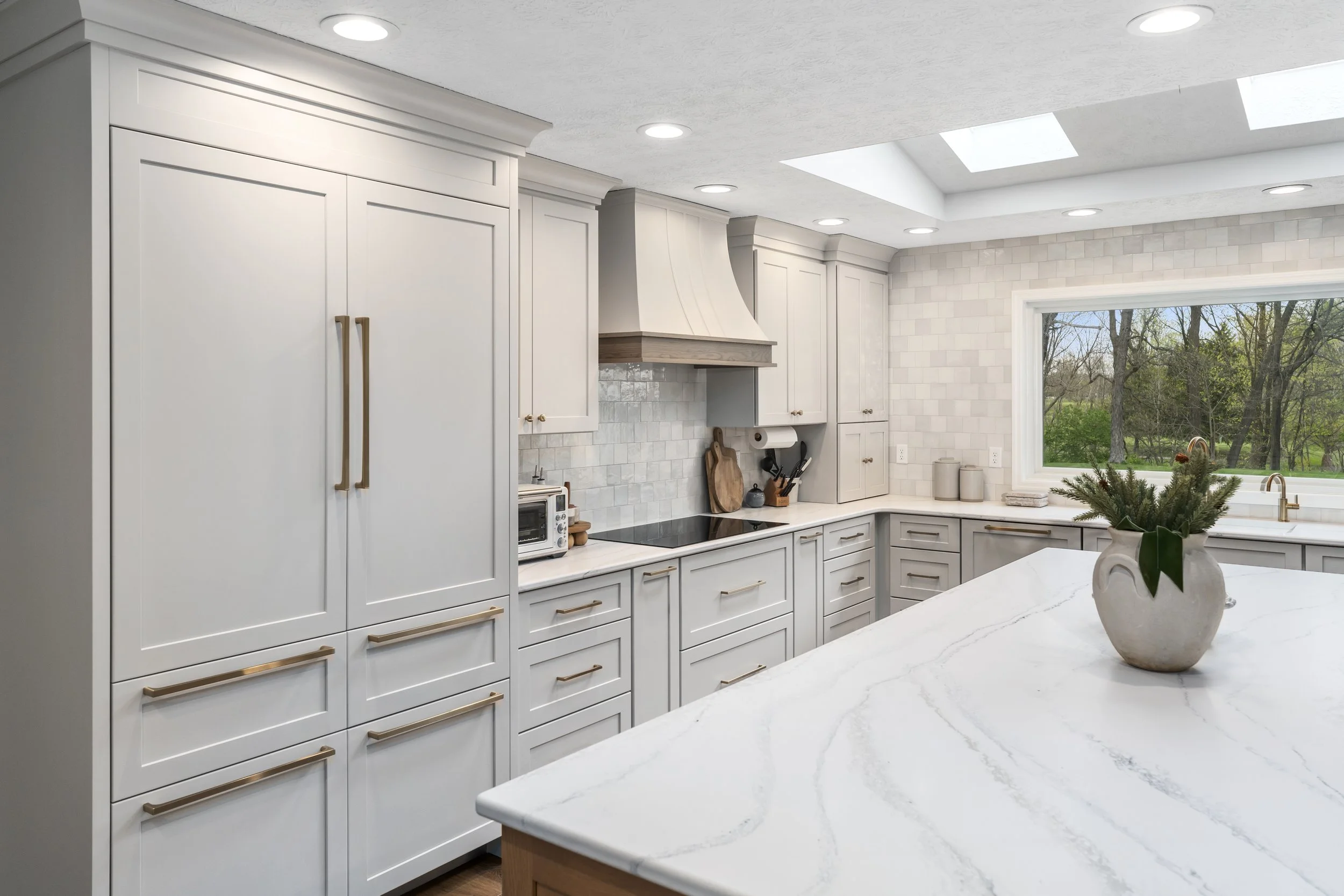 Renovated kitchen with white cabinetry, brass hardware, tile backsplash, integrated cooktop, and a large window above the counter for natural light