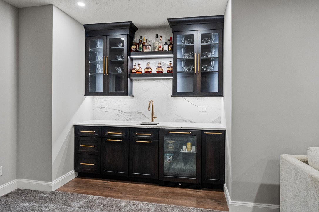 Basement wet bar with dark glass-front cabinets, backlit display shelving, and a marble backsplash in a Brookville, Ohio basement renovation.