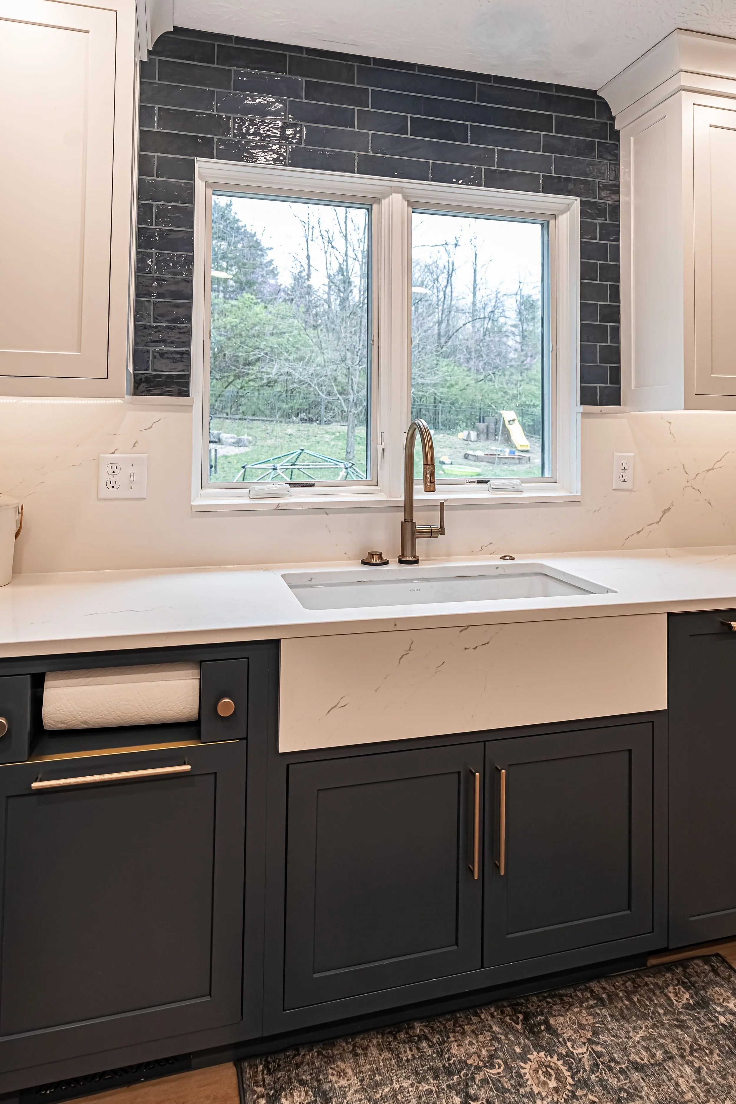 Apron-front farmhouse sink with a brass faucet centered under a window above navy cabinetry in a Centerville kitchen.