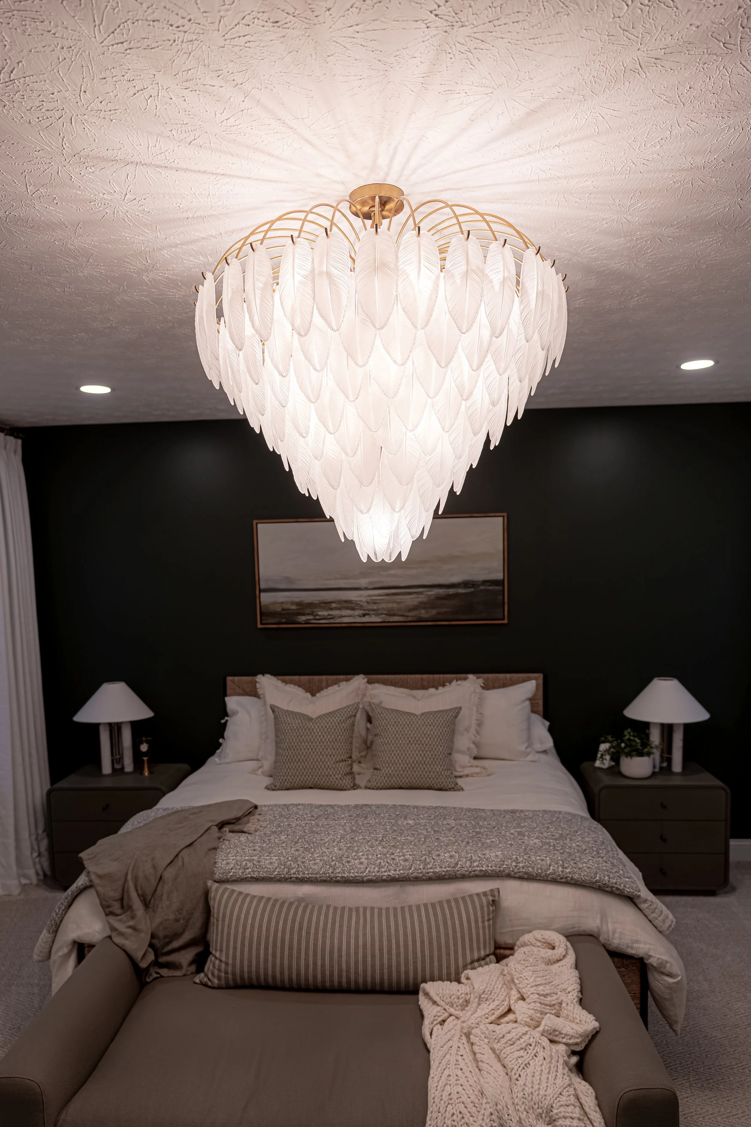 Master bedroom with a sculptural feather chandelier, dark accent wall, symmetrical nightstands and lamps, and layered bedding in an Oakwood, Ohio renovation