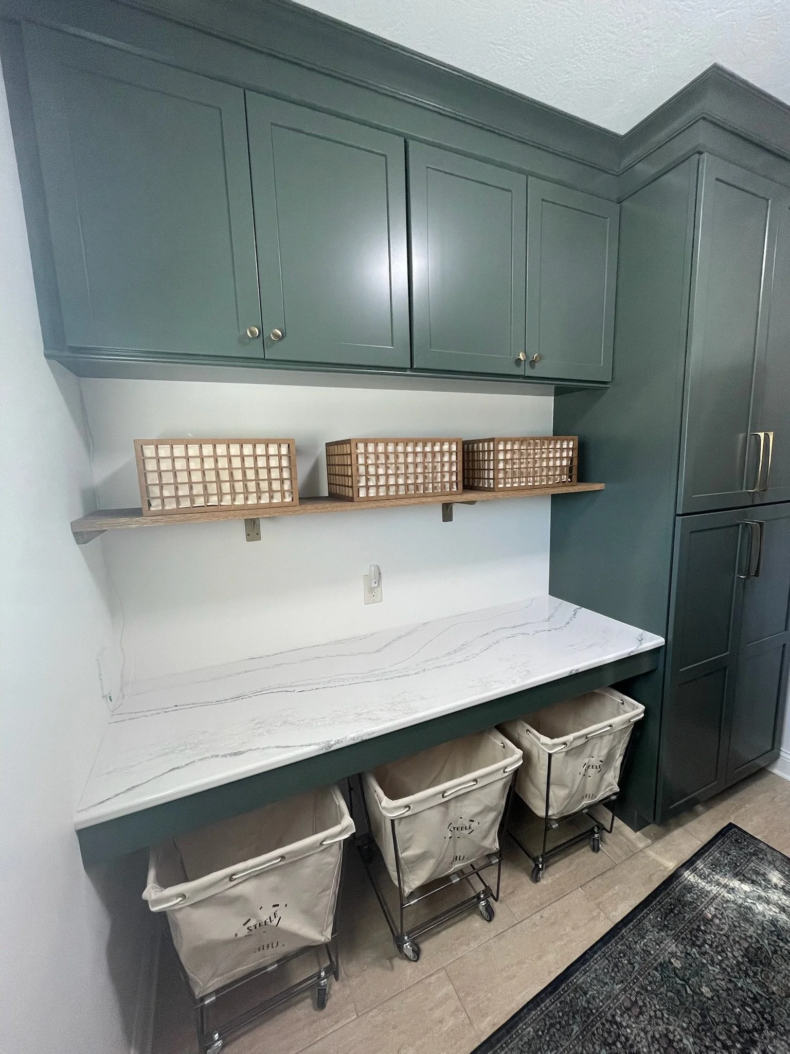 Laundry folding station with green cabinetry, a quartz counter, wood shelf with woven baskets, and three rolling pull-out hampers in a Beavercreek, Ohio home