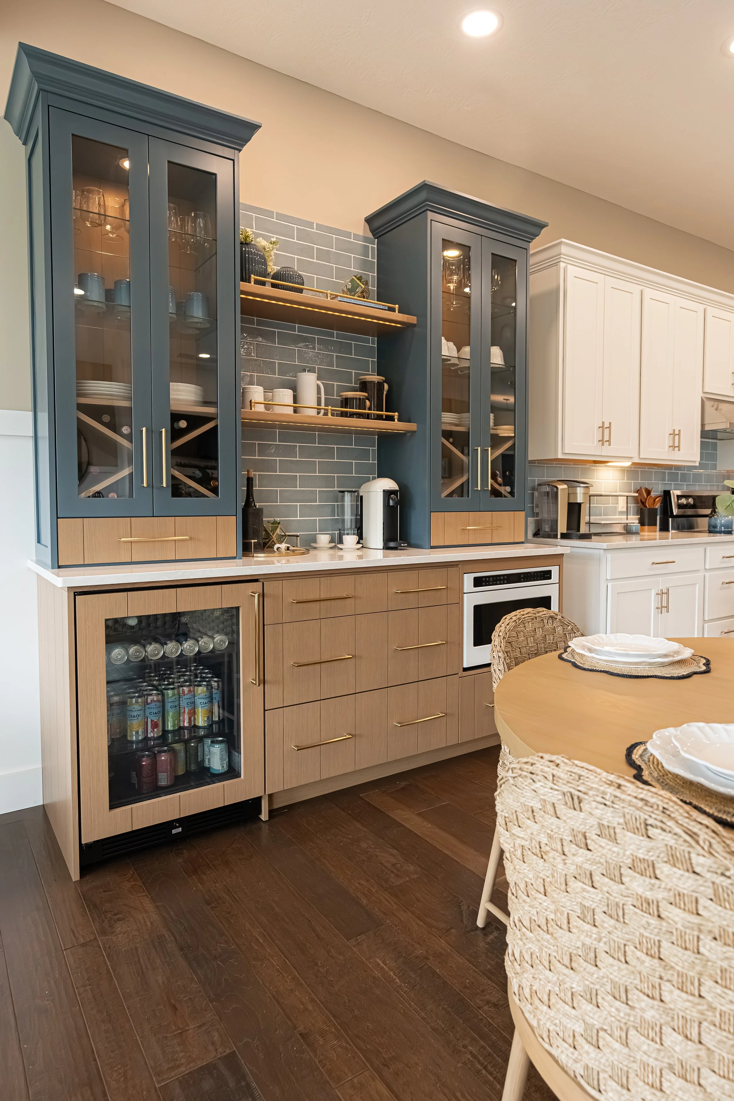 A kitchen with blue and white cabinets, open shelves, and a countertop with a coffee maker, cups, and small decor. The background has a gray tile backsplash and dark wood flooring.
