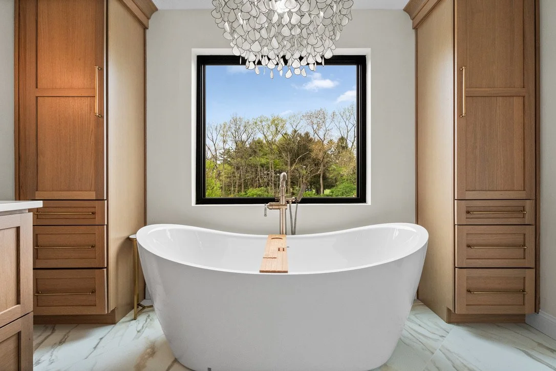 Master bathroom with a freestanding soaking tub, crystal chandelier, and brass tub filler flanked by wood storage towers in a Brookville, Ohio renovation.