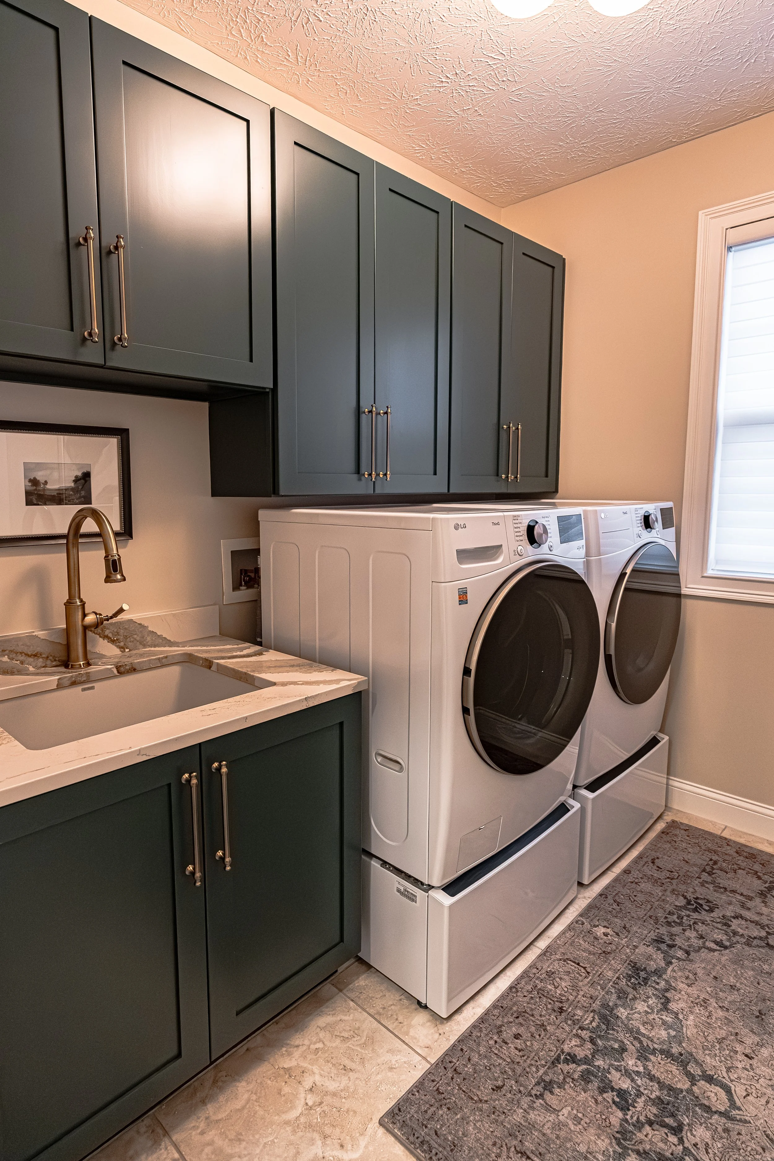 Renovated laundry room with deep green cabinetry, brass hardware, a stone countertop and sink, brass faucet, and front-load washer and dryer in an Oakwood, Ohio home