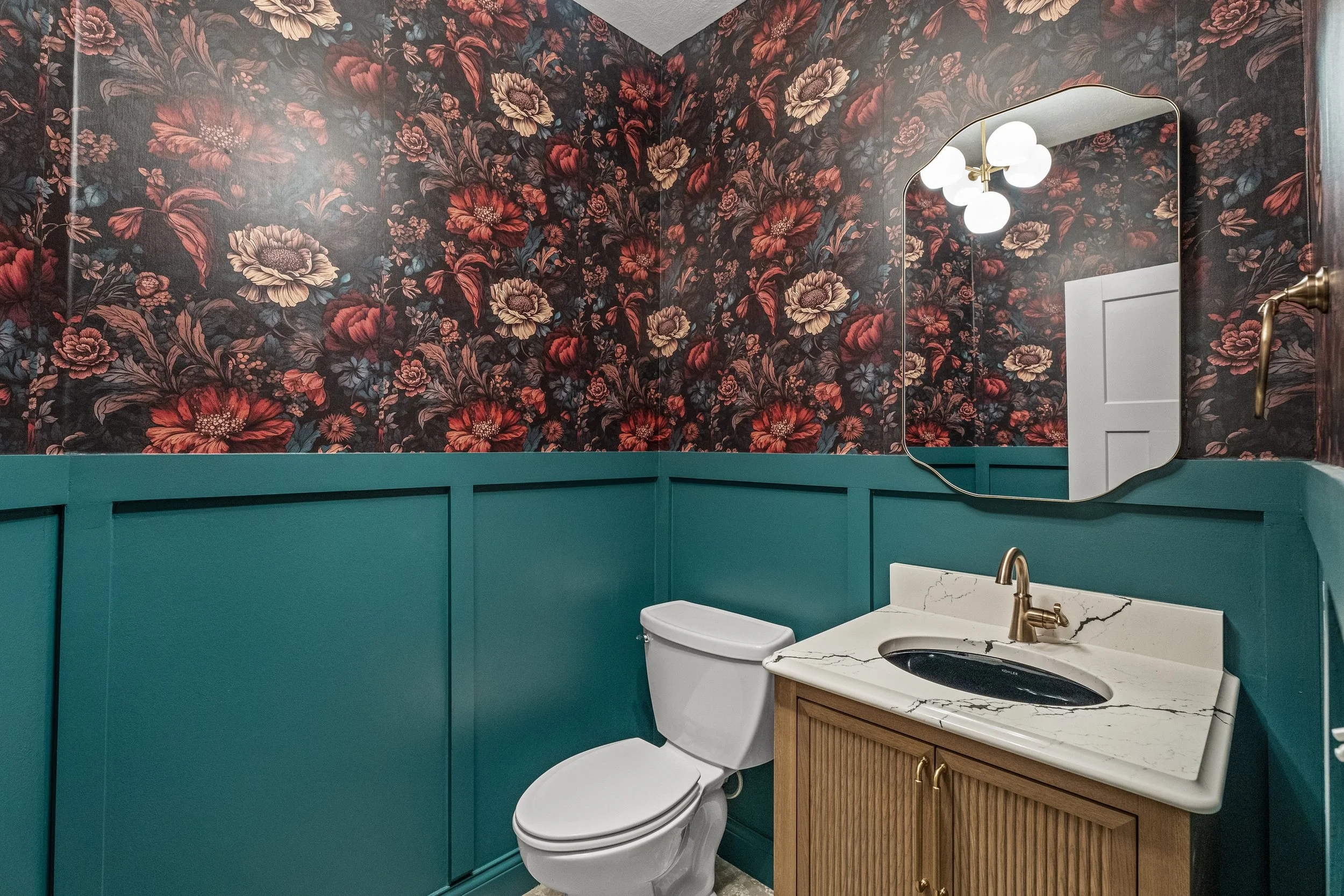Basement half bath with dark floral wallpaper, deep green wainscoting, a white oak vanity with marble-look countertop, and brass fixtures