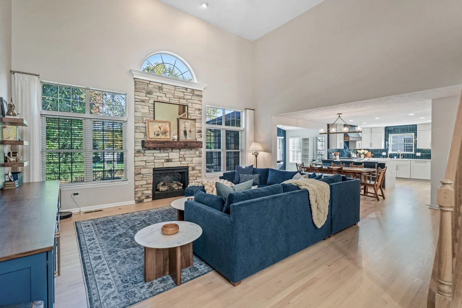 Open-concept living room with a stone fireplace, blue sectional, and vaulted ceiling flowing to the kitchen in a Beavercreek, Ohio main-level remodel.