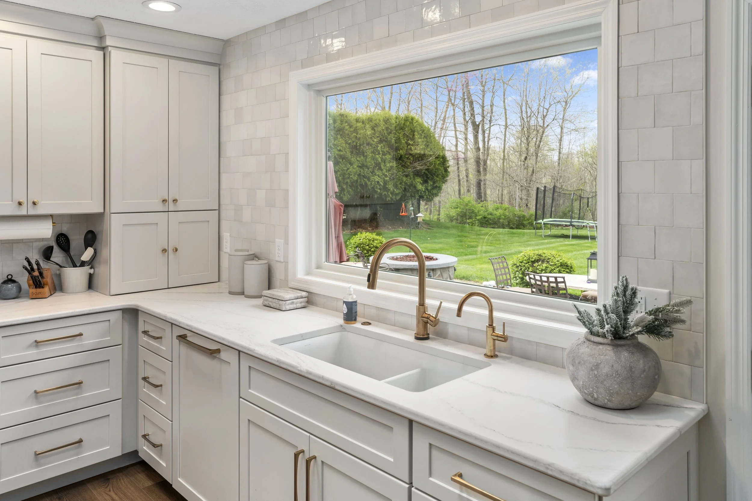 Kitchen sink with brass faucet beneath a large new window overlooking the backyard, with white cabinetry and quartz countertops