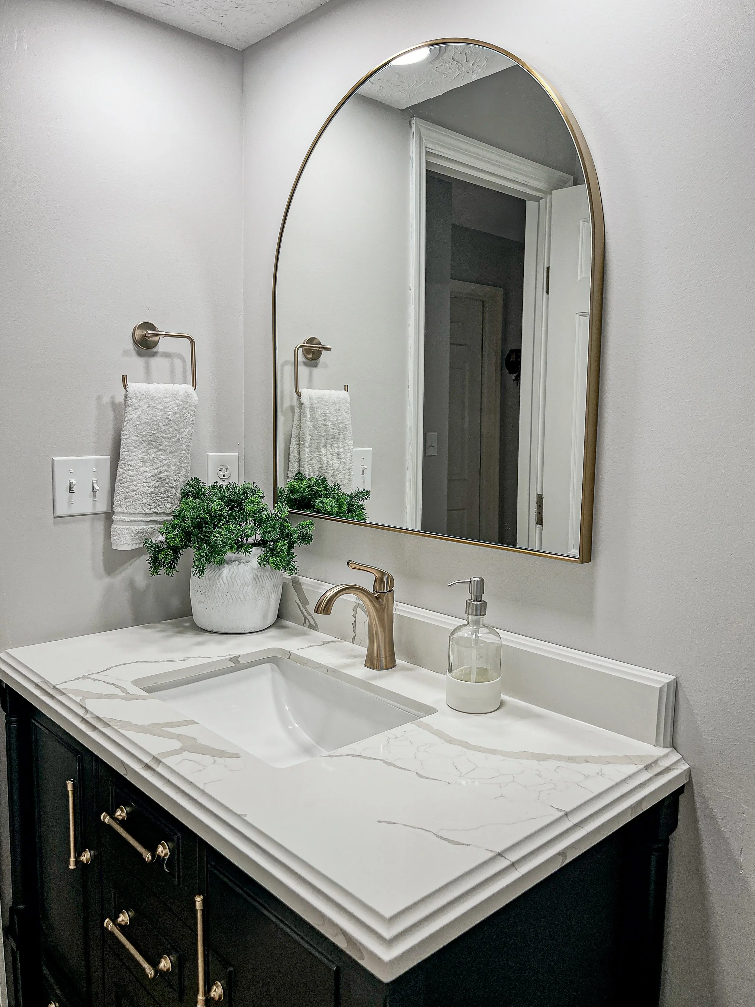 Bathroom with a dark vanity, quartz countertop, brass faucet, and gold arched mirror in a Centerville whole-home remodel.