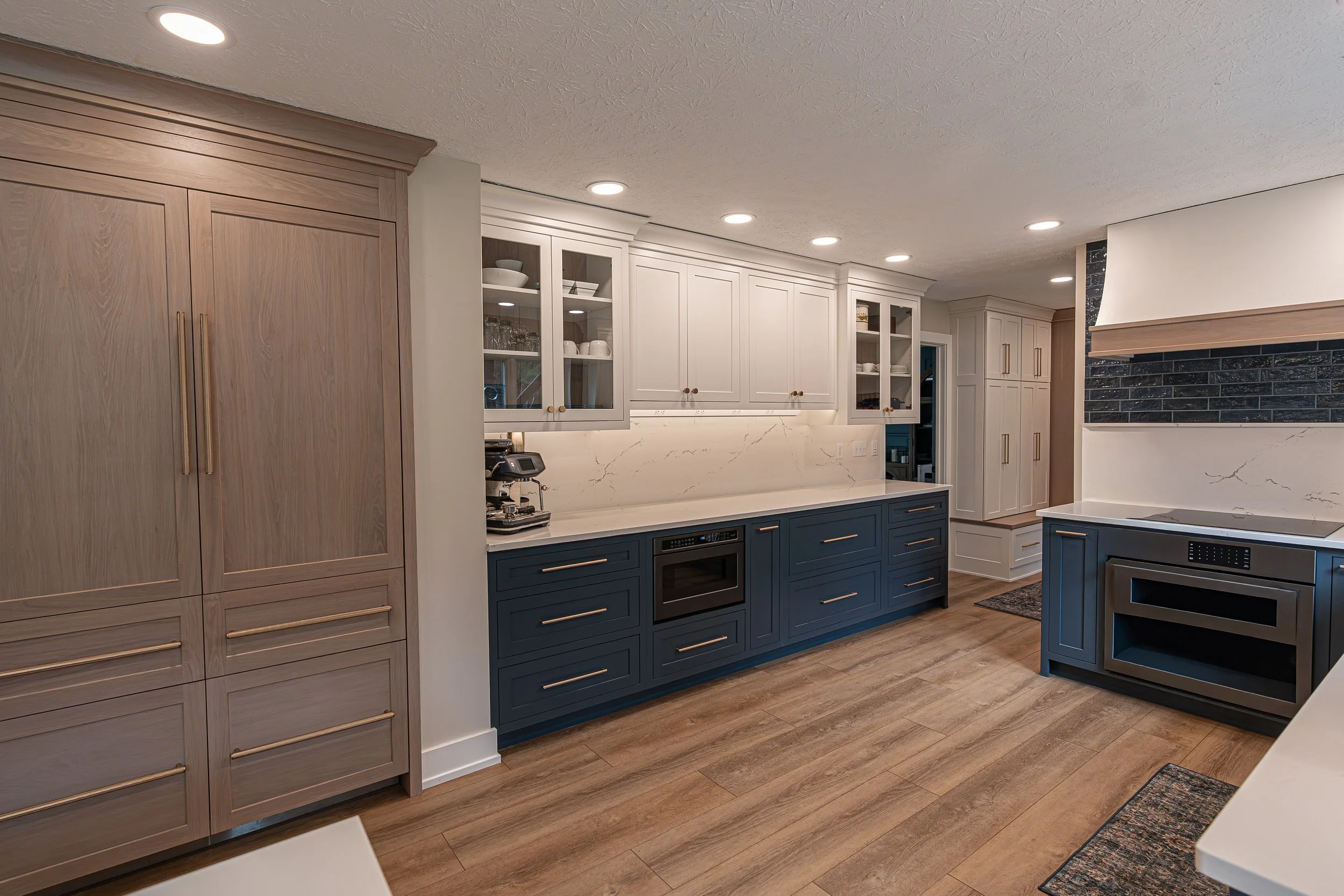 Modern kitchen with beige and dark blue cabinets, white marble countertops, glass-front cabinets, wood flooring, and recessed ceiling lights.